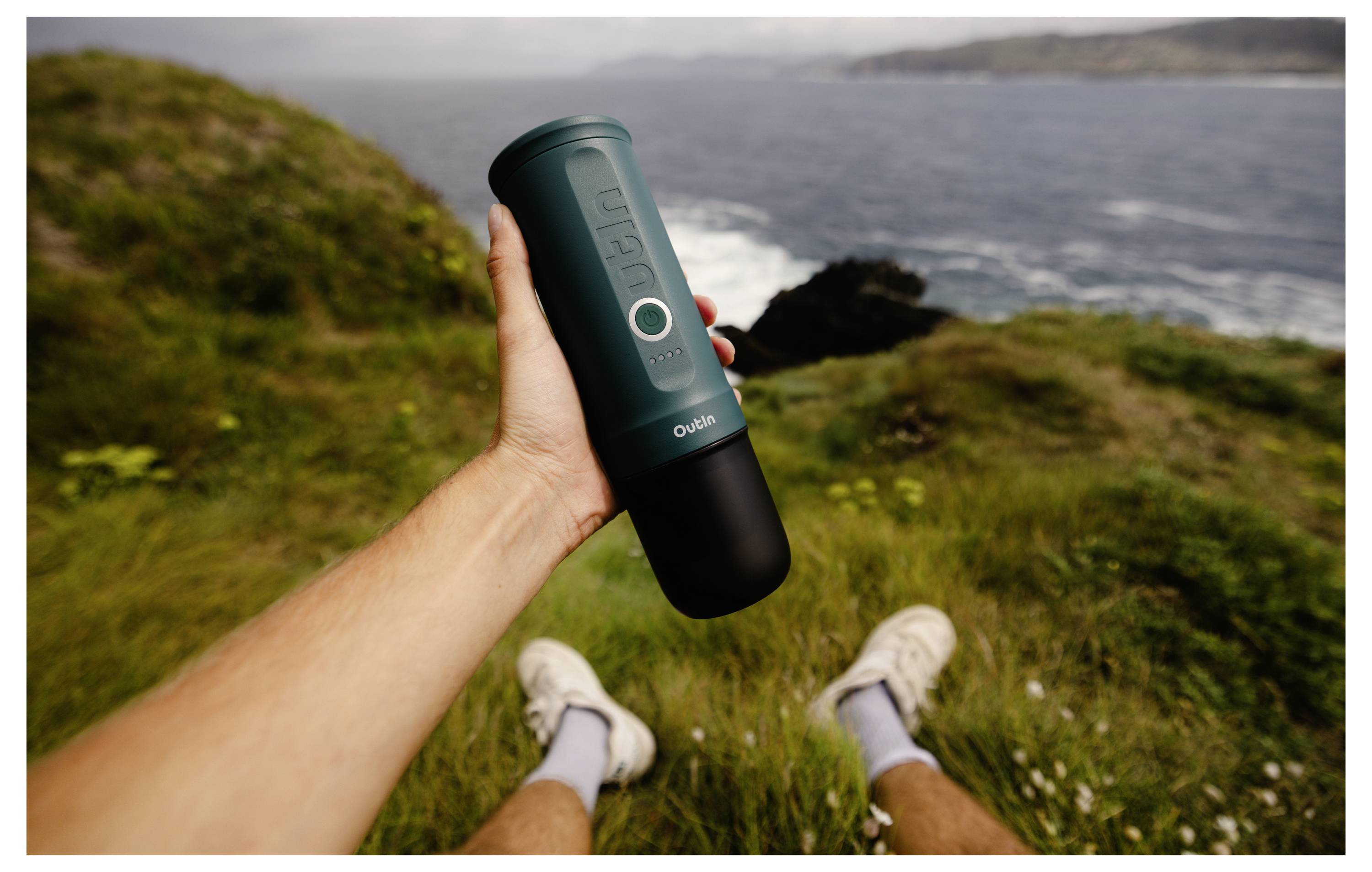 A person holds a portable coffee maker on a grassy cliff overlooking the ocean, featuring a distant view of coastlines under a cloudy sky.