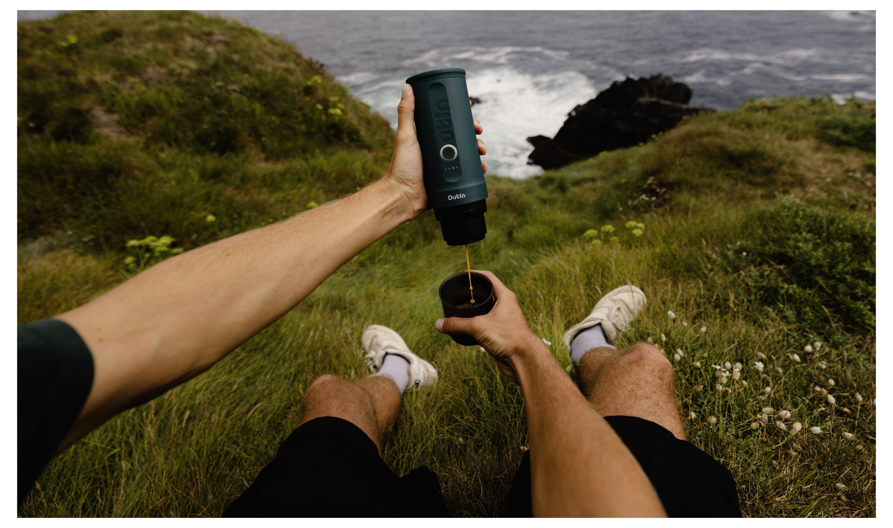 A person sitting on grass near the ocean pours coffee from a portable coffee maker into a cup, with cliffs and waves in the background.