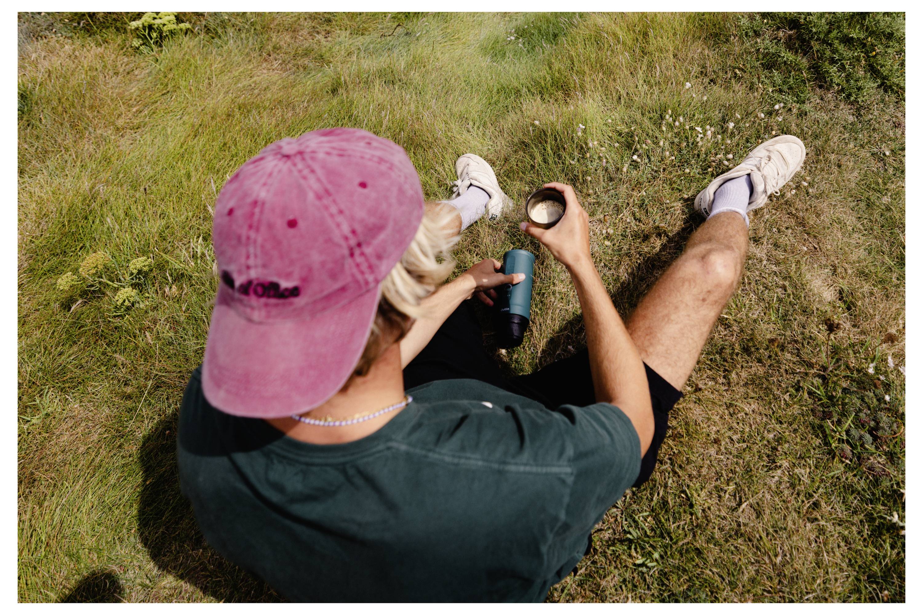 A person sitting on grass, wearing a red cap and green shirt, holding a thermos and cup, likely relaxing or enjoying a drink outdoors.