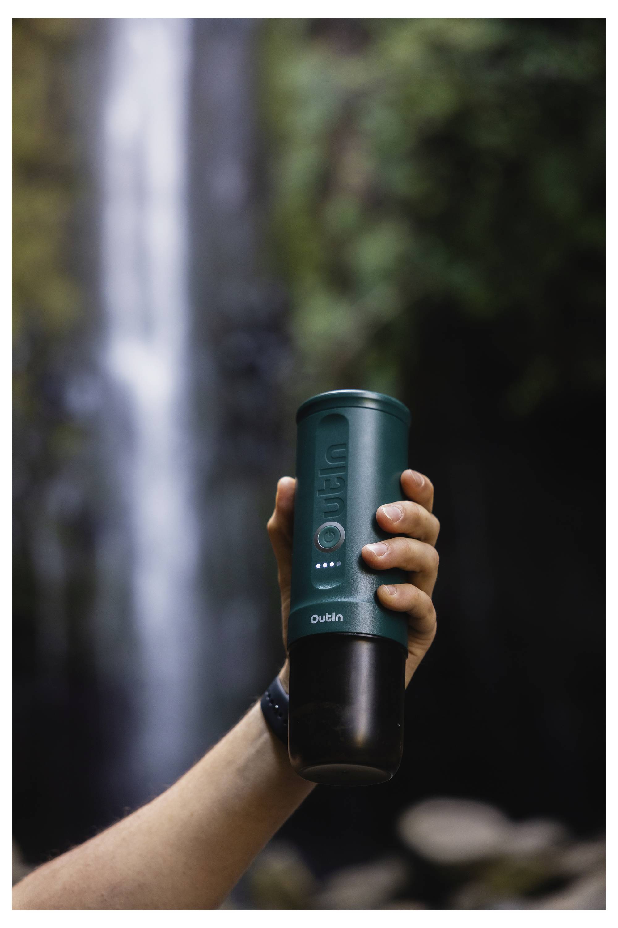 A hand holds a green portable coffee maker with a waterfall in the blurred background, depicting outdoor use in a natural setting.