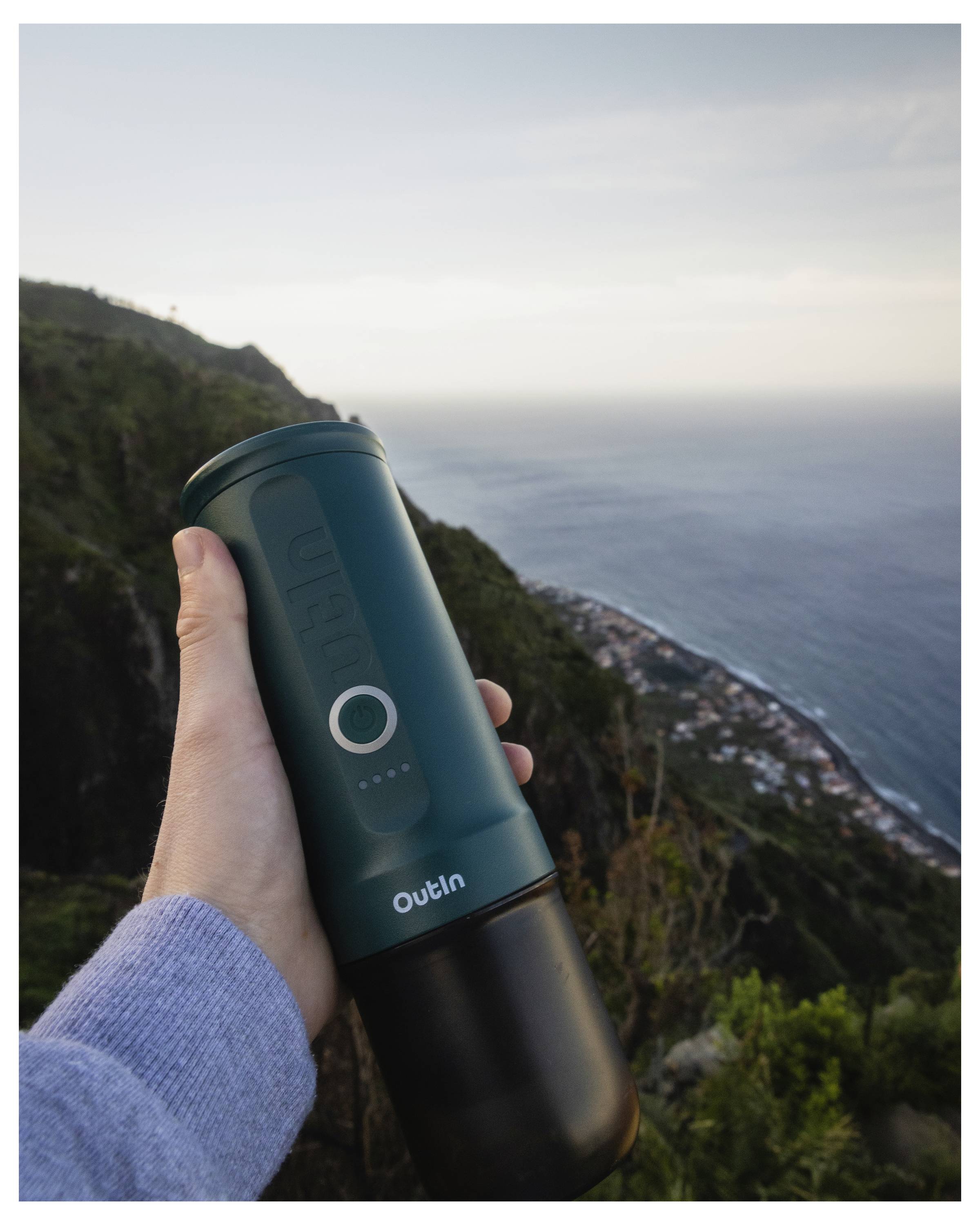 A hand holds a green portable coffee grinder with hills and the ocean in the background. The grinder shows a circular light indicator.