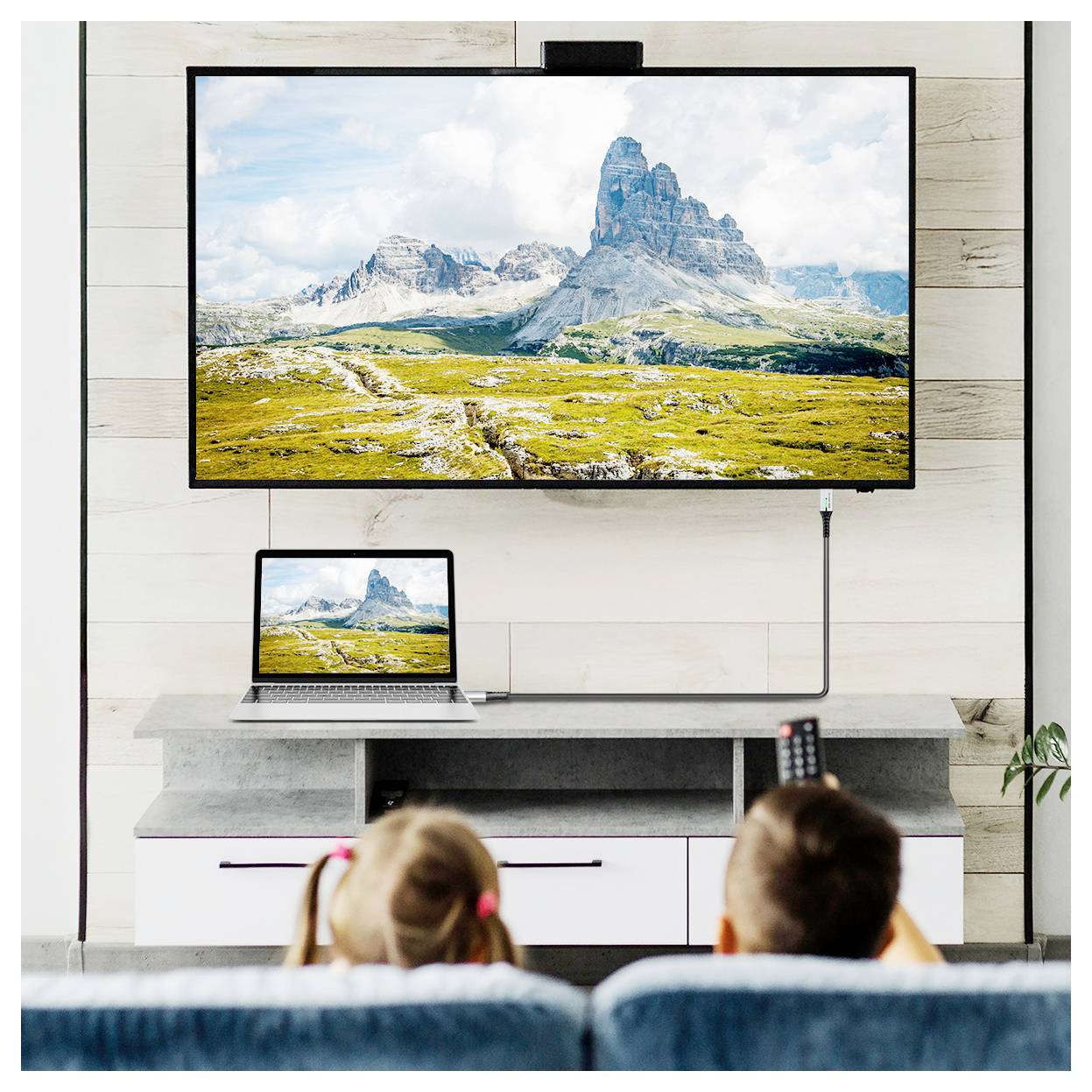 Two children sit on a couch watching a large wall-mounted TV displaying a scenic landscape. A laptop with the same image is on a table below.
