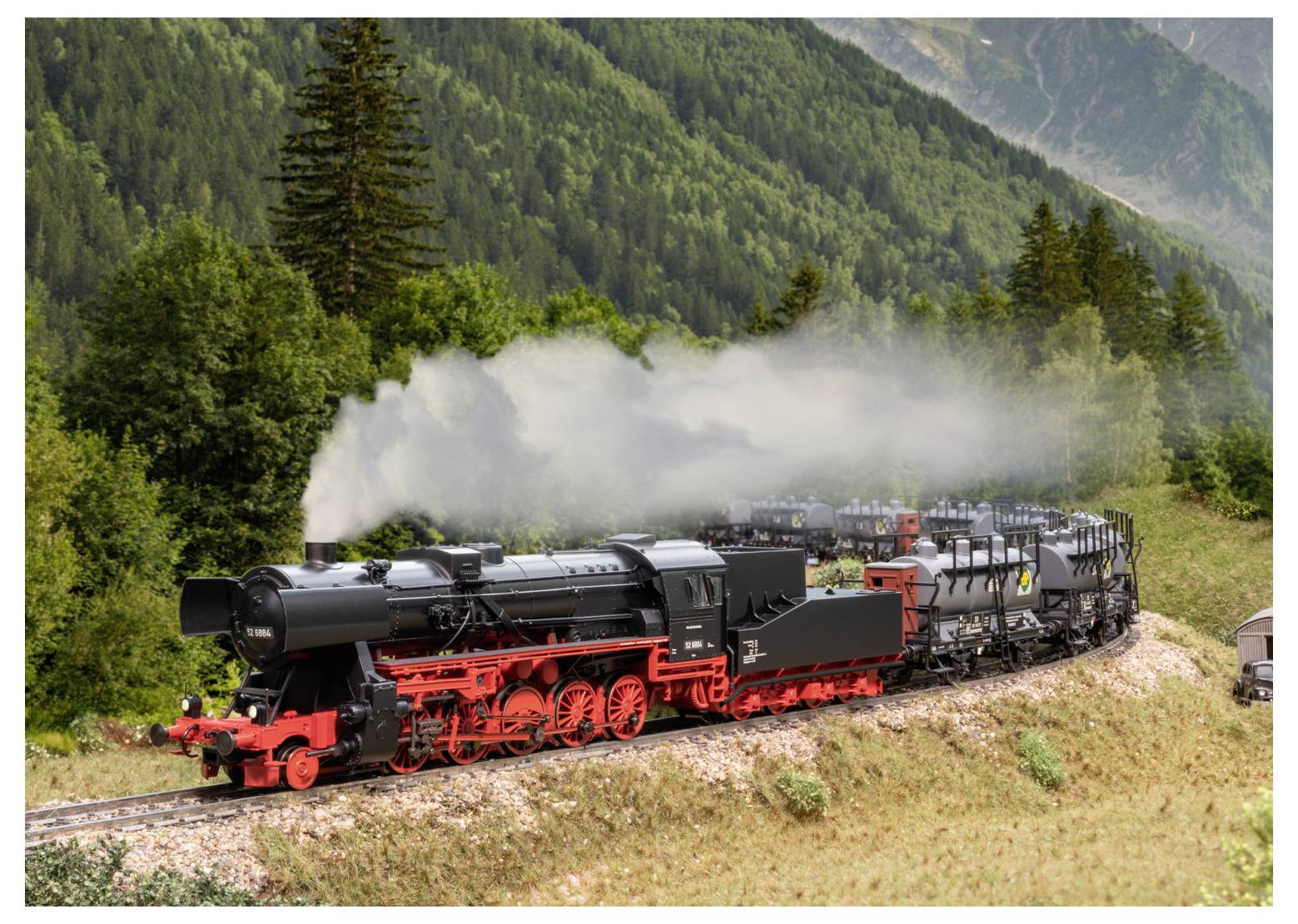 A steam locomotive with a red front travels on a track through a lush green mountainous landscape, emitting white smoke.