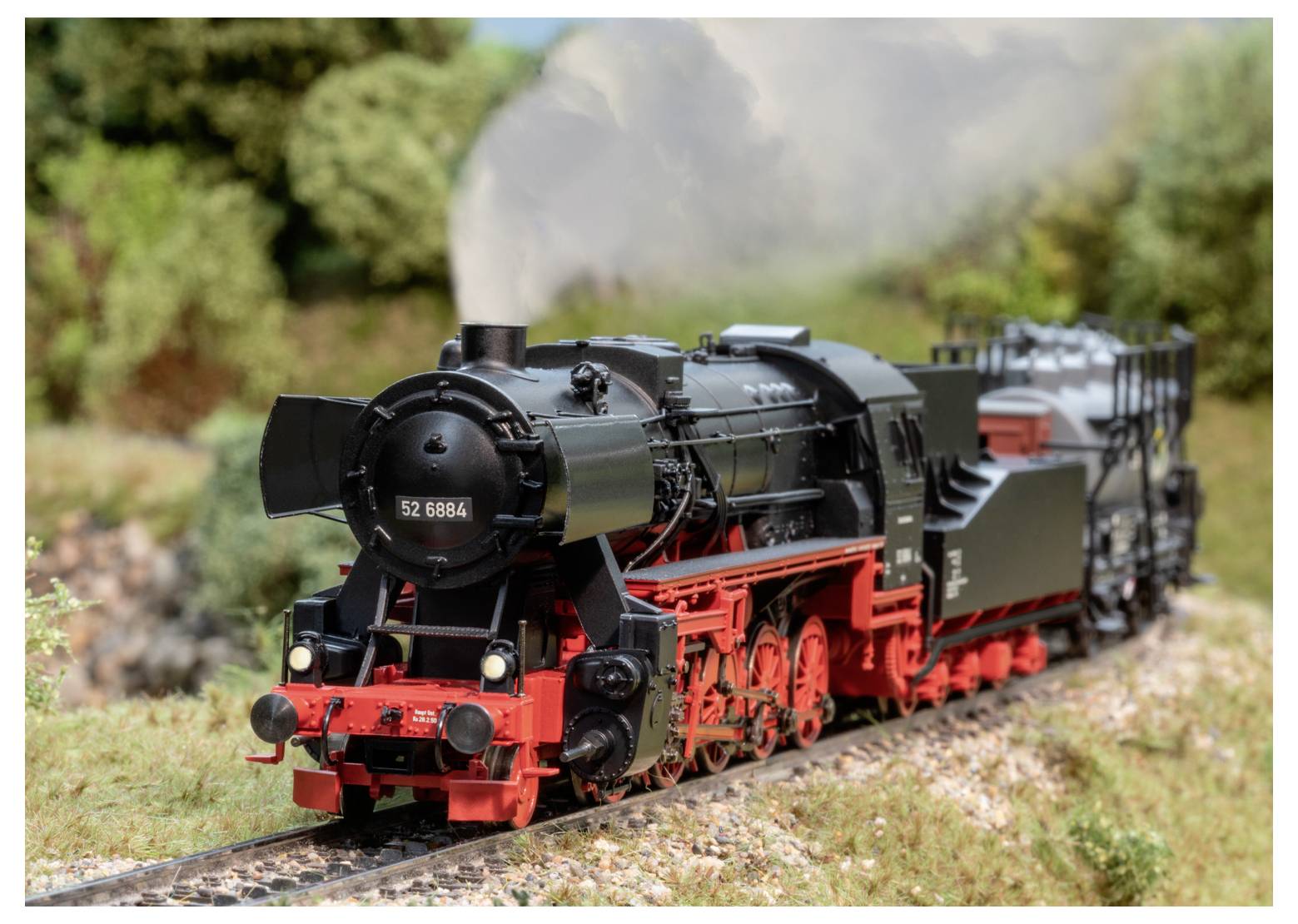 A black and red steam locomotive travels on a model railway track through a green landscape, emitting white smoke from its chimney.