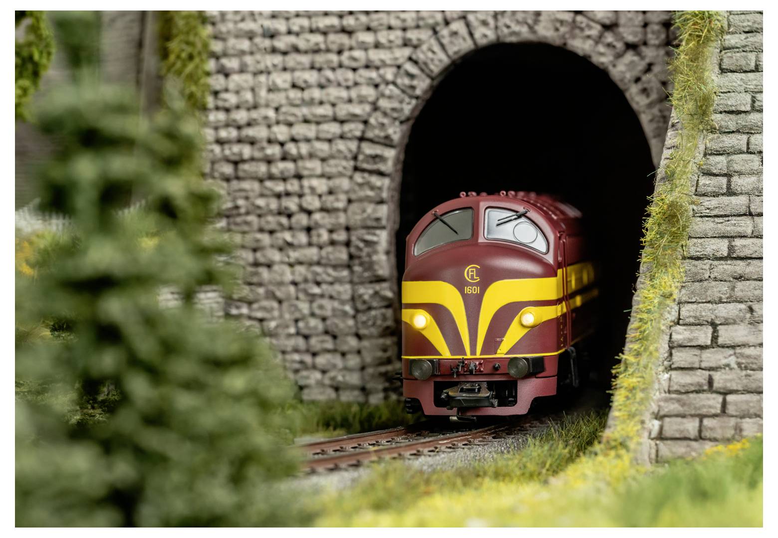 A vintage-style train, maroon with yellow stripes, emerges from a stone tunnel surrounded by greenery and grass.