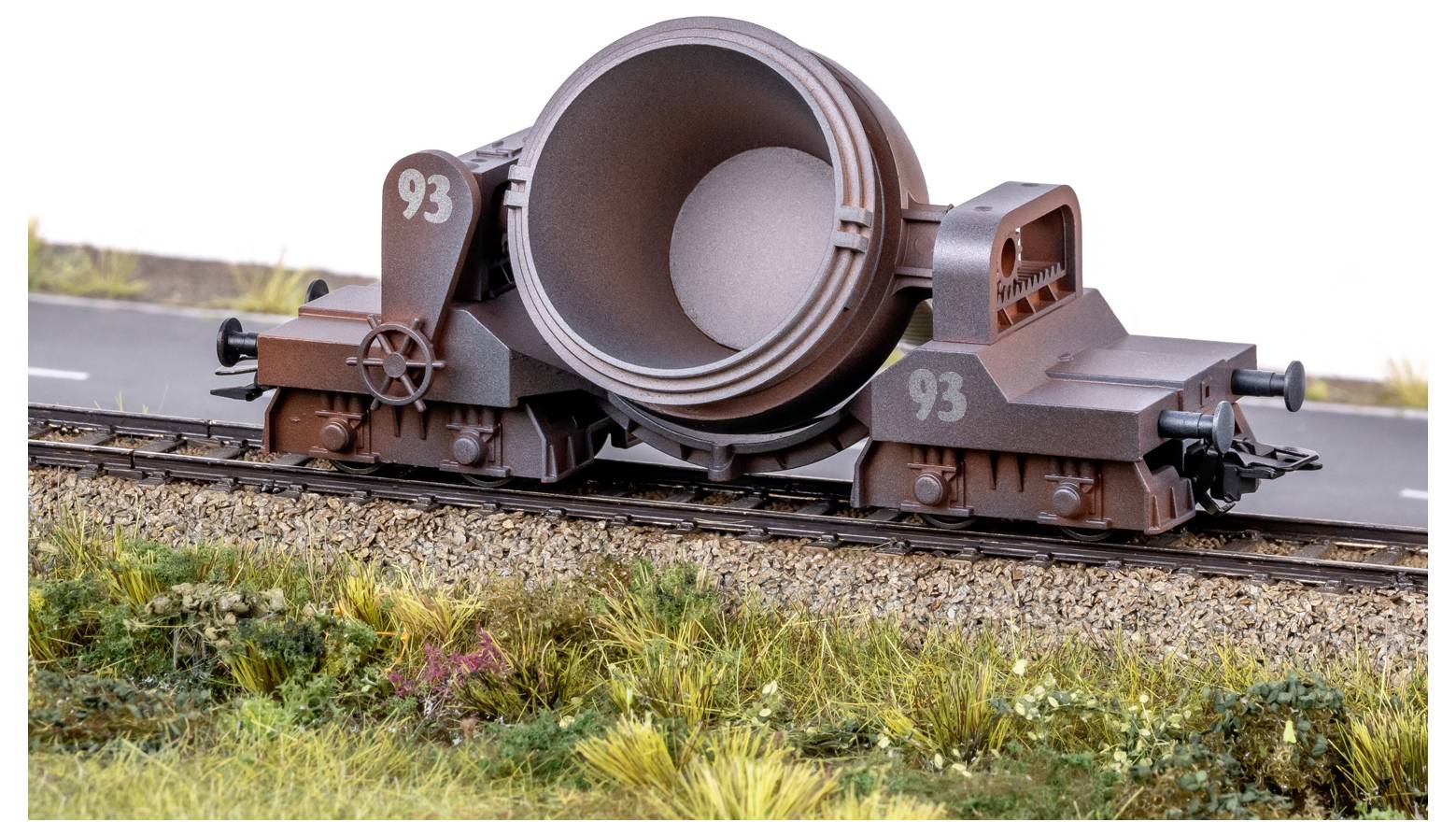 A model of a brown industrial rail car with a large cylindrical container labeled '93', set on train tracks with grass and road nearby.