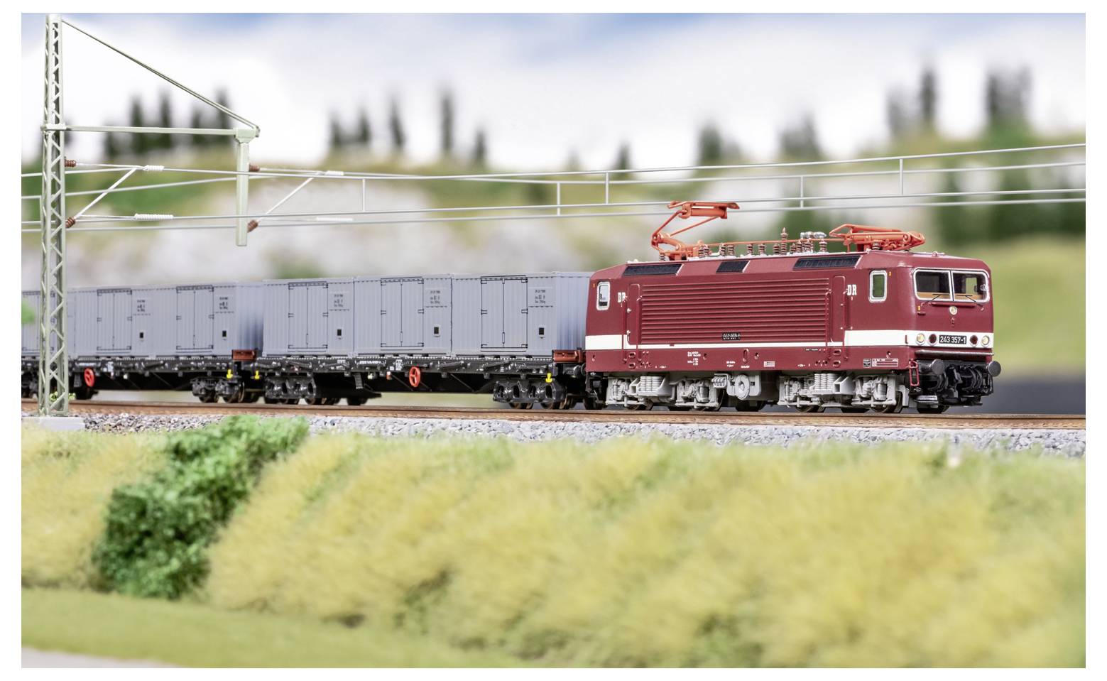 Red electric freight locomotive pulling gray cargo cars on a model railway track under overhead power lines.