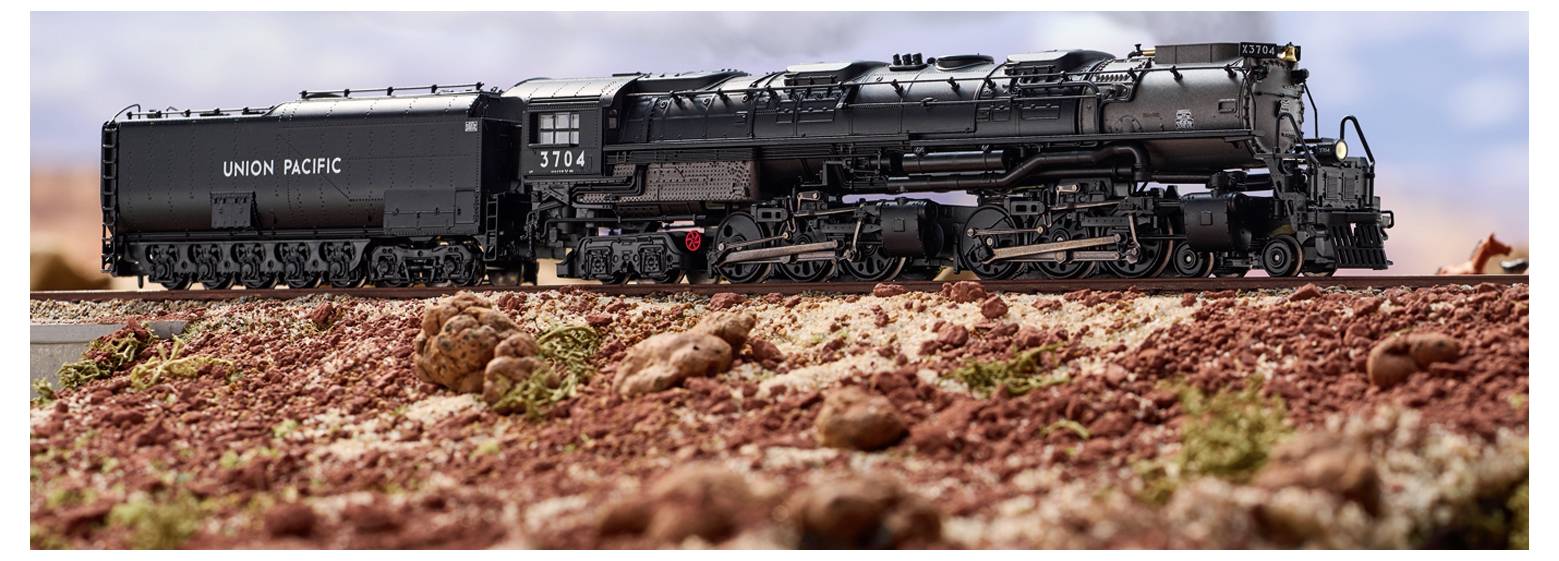 A black Union Pacific steam locomotive numbered 3704 is displayed on a track against a rocky landscape with a cloudy sky in the background.