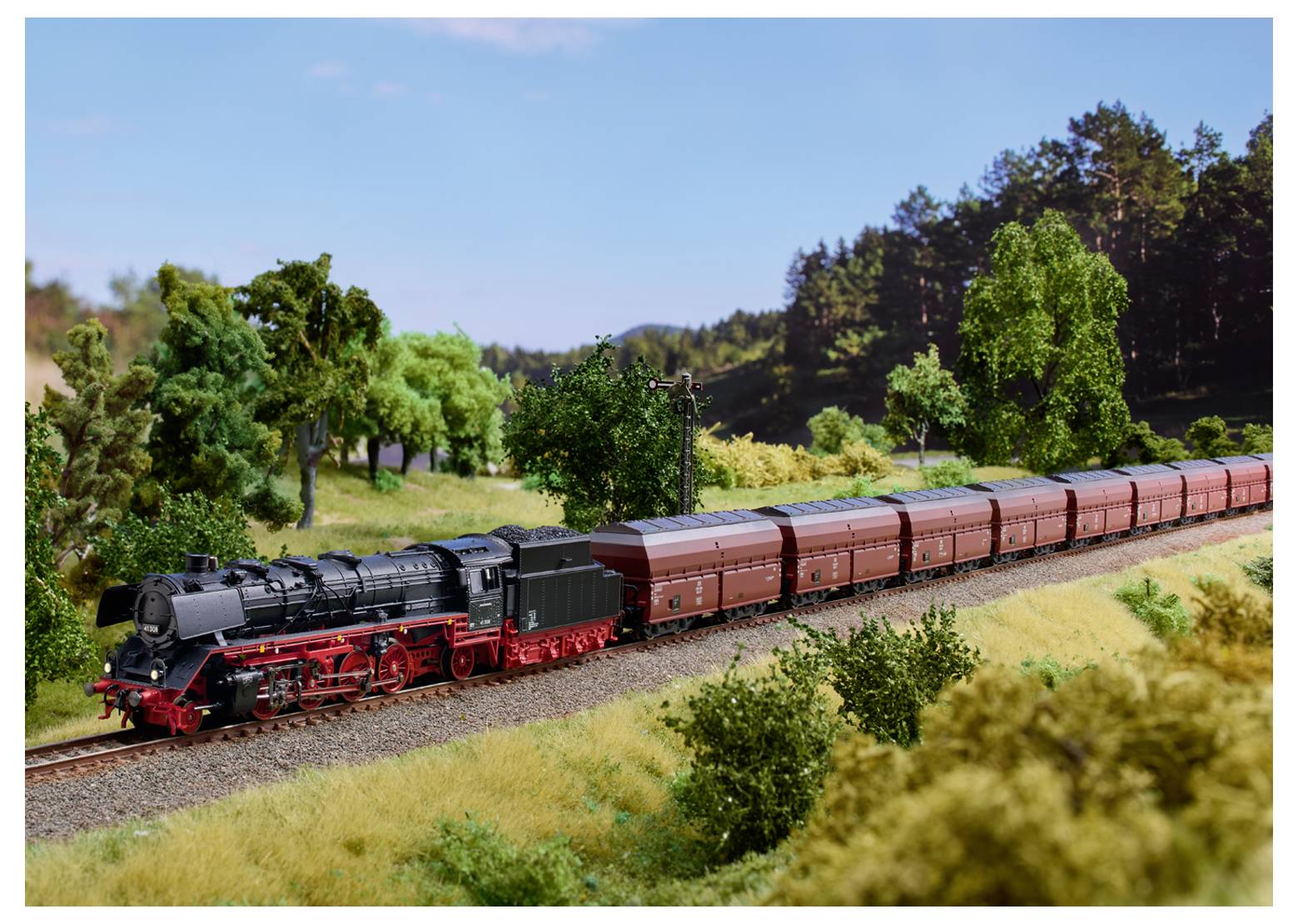 A model train with red and black steam engine pulls brown freight cars through a scenic, grassy landscape with trees in the background.