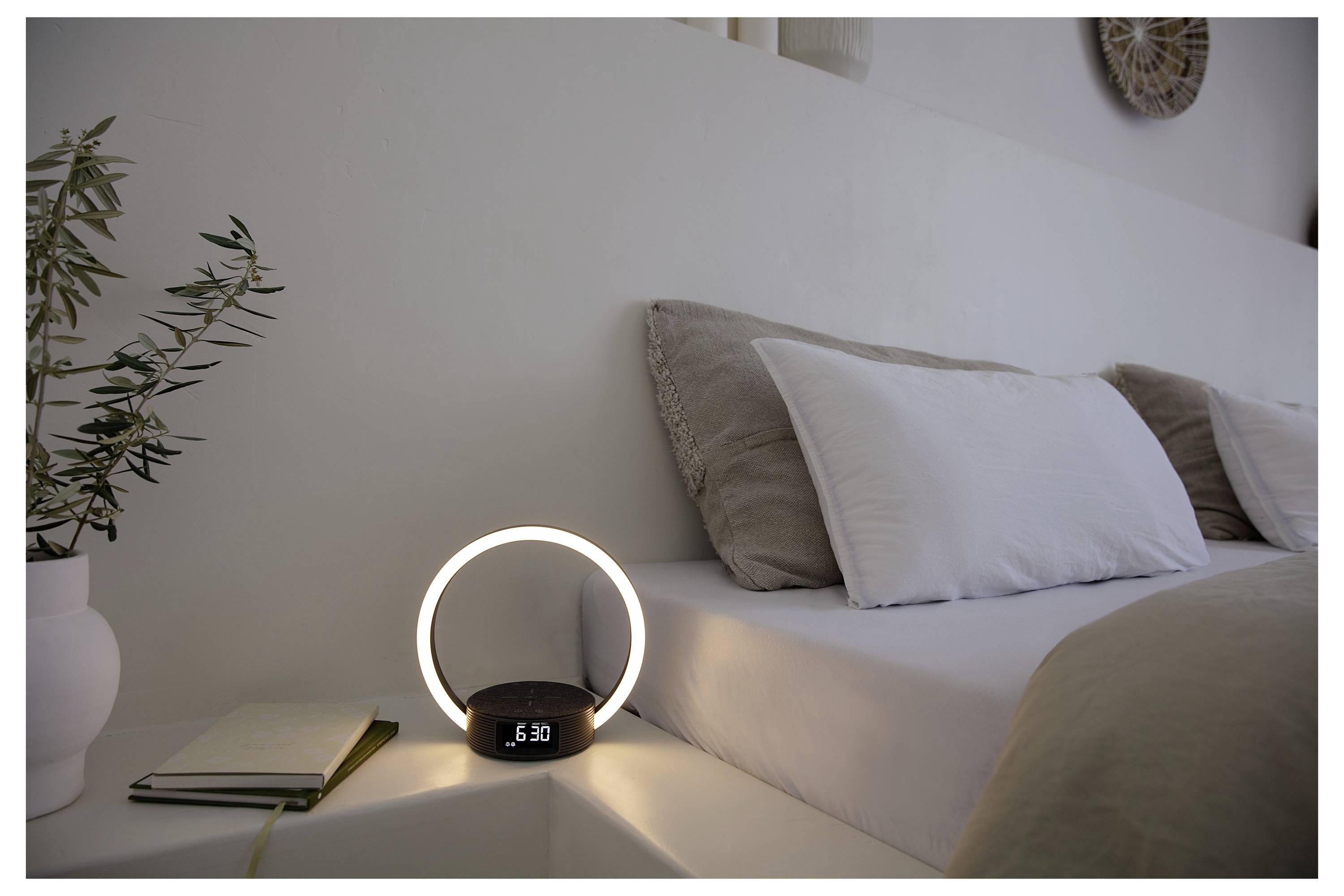 A neatly made bed with white linens in a bright room. A glowing ring-shaped lamp showing '5:30' on a nightstand, beside a notebook and plant.