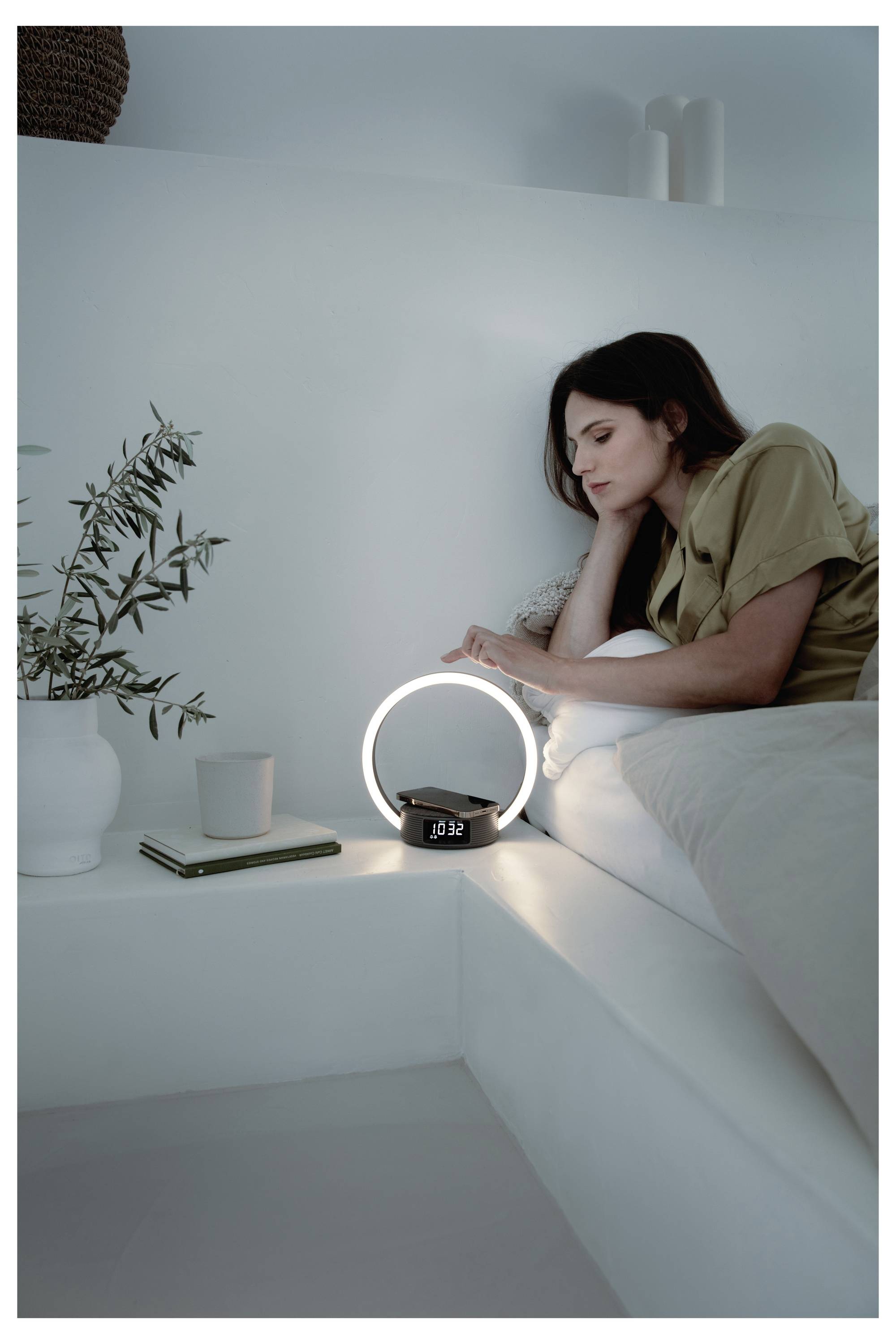 A woman in a beige outfit sits on a bed, leaning on her elbow, gazing at a glowing circular lamp beside a digital clock displaying '10:38'.