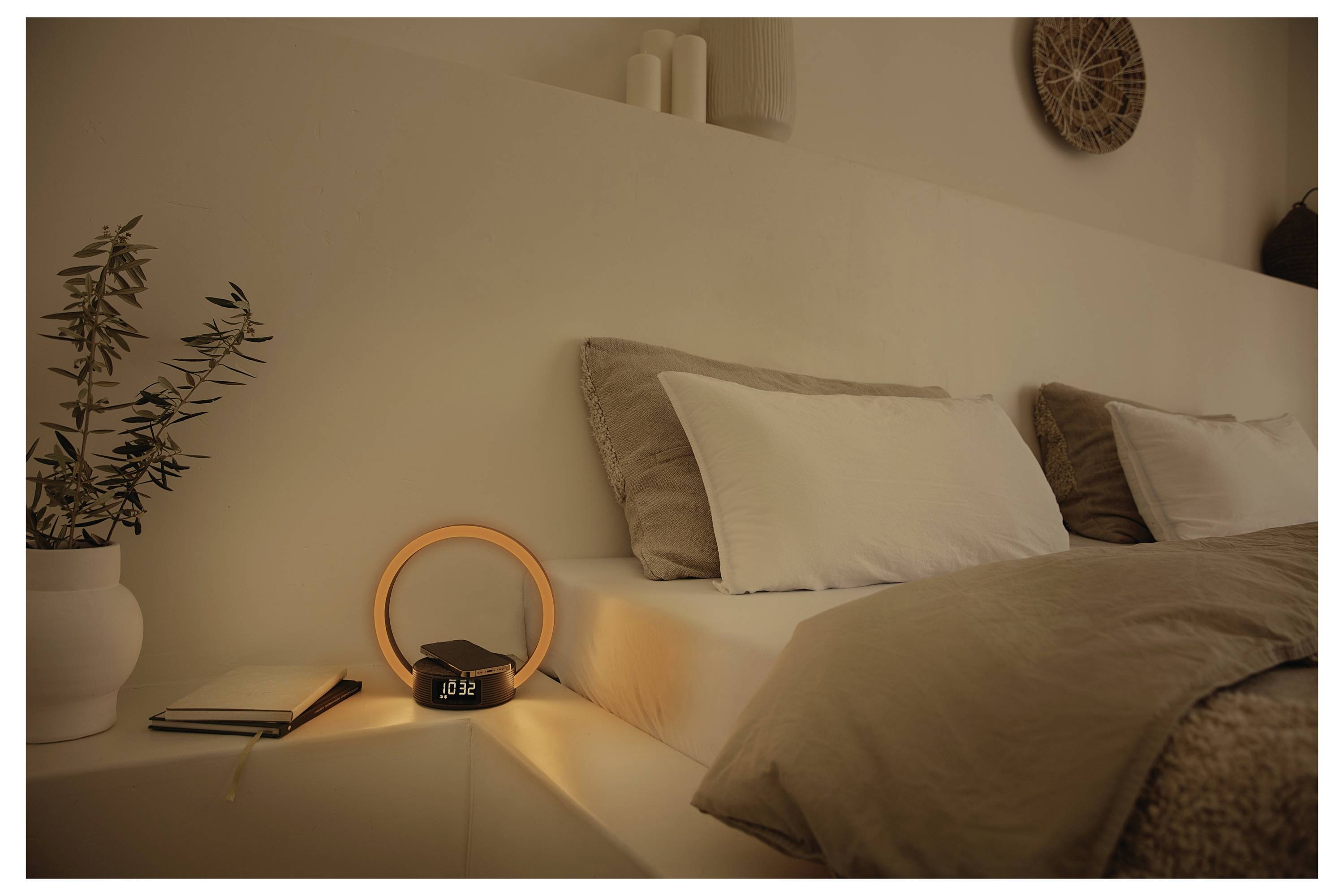 'A cozy bedroom with neutral bedding, warm lighting from a ring-shaped lamp on a nightstand, beside a small clock showing 10:12.'