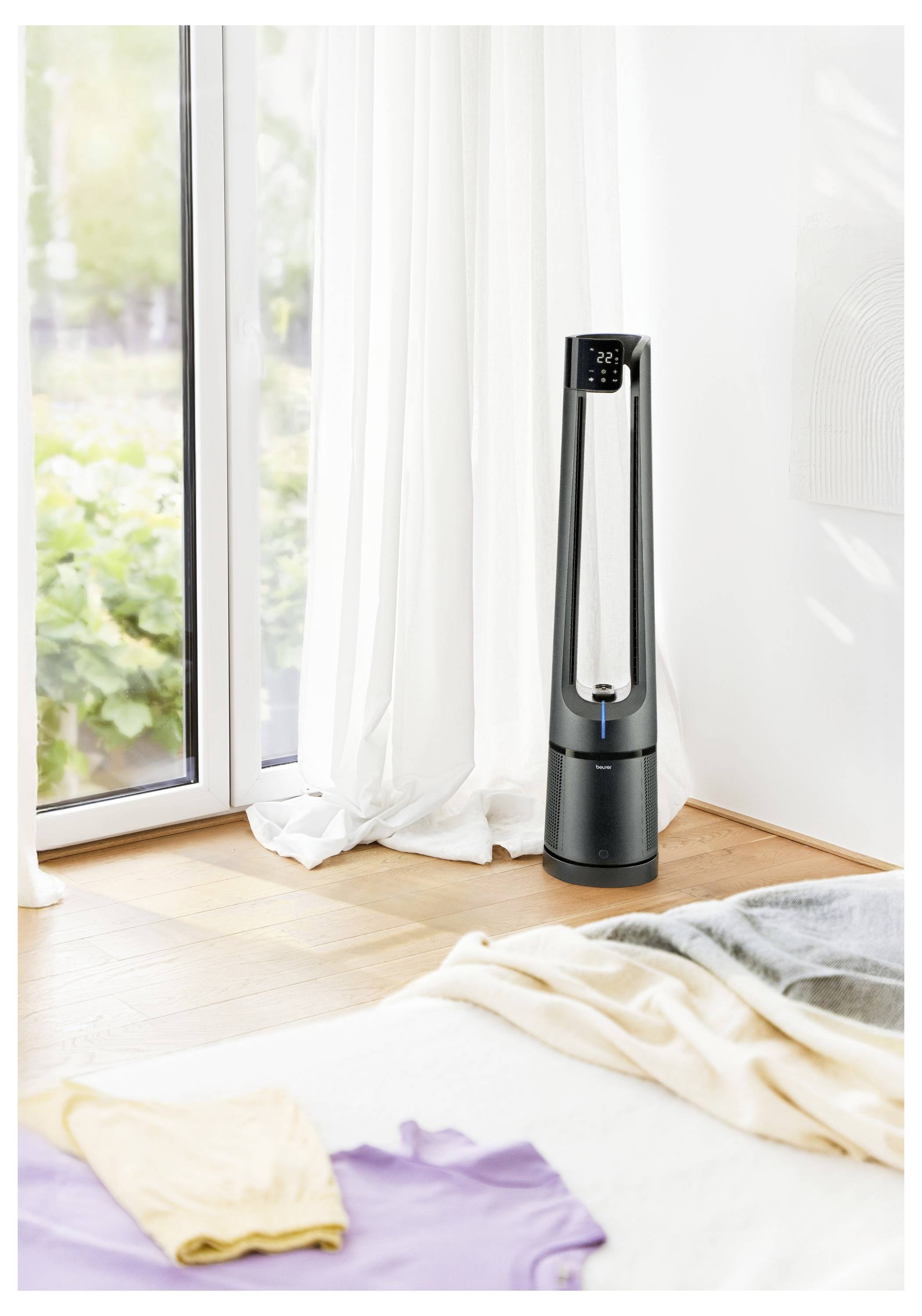 'Tower fan in modern room by large window, with sunlight filtering in and laundry on bed.'