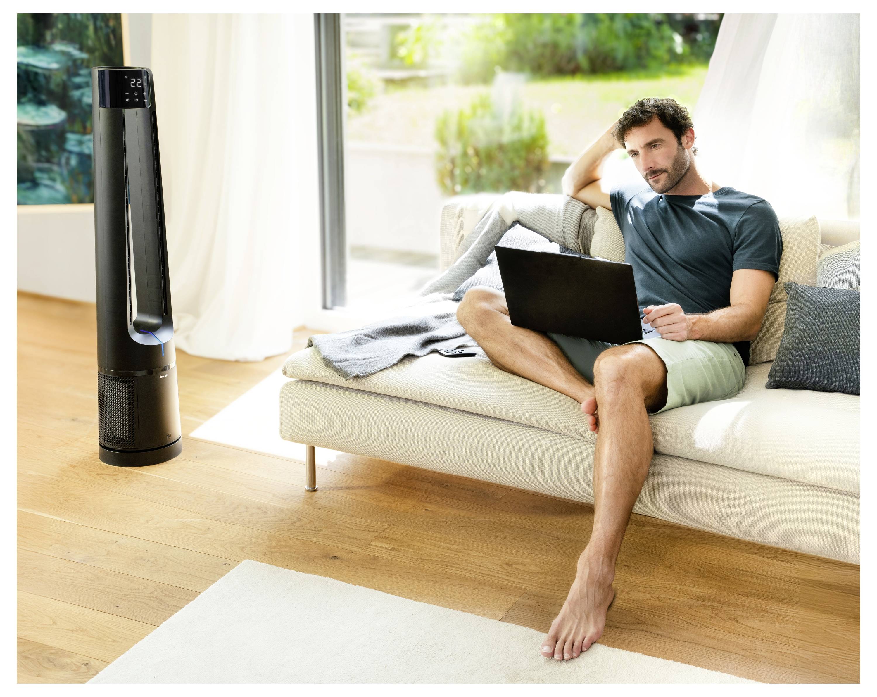 A man relaxes on a couch, using a laptop, with a tall, modern fan nearby. Bright, airy room with large windows and greenery outside.