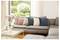 A cozy living room with a grey sectional sofa topped with colorful cushions in pink, peach, teal, and beige. A vase with pink flowers sits on a white table, and a large window reveals a garden outside.