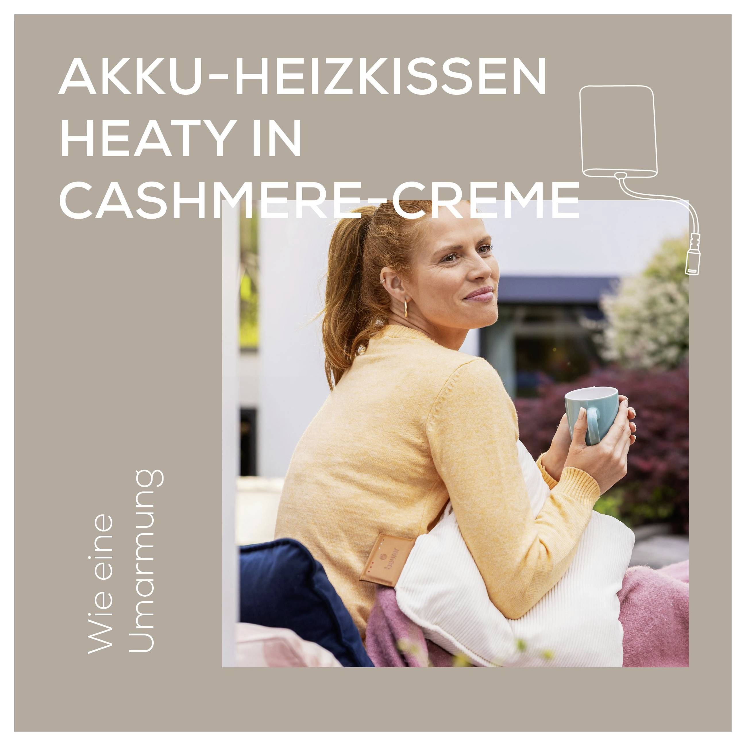 A woman in a peach sweater sits outside on a patio chair, holding a blue mug and a heated cushion labeled 'AKKU-HEIZKISSEN HEATY IN CASHMERE-CREME'.