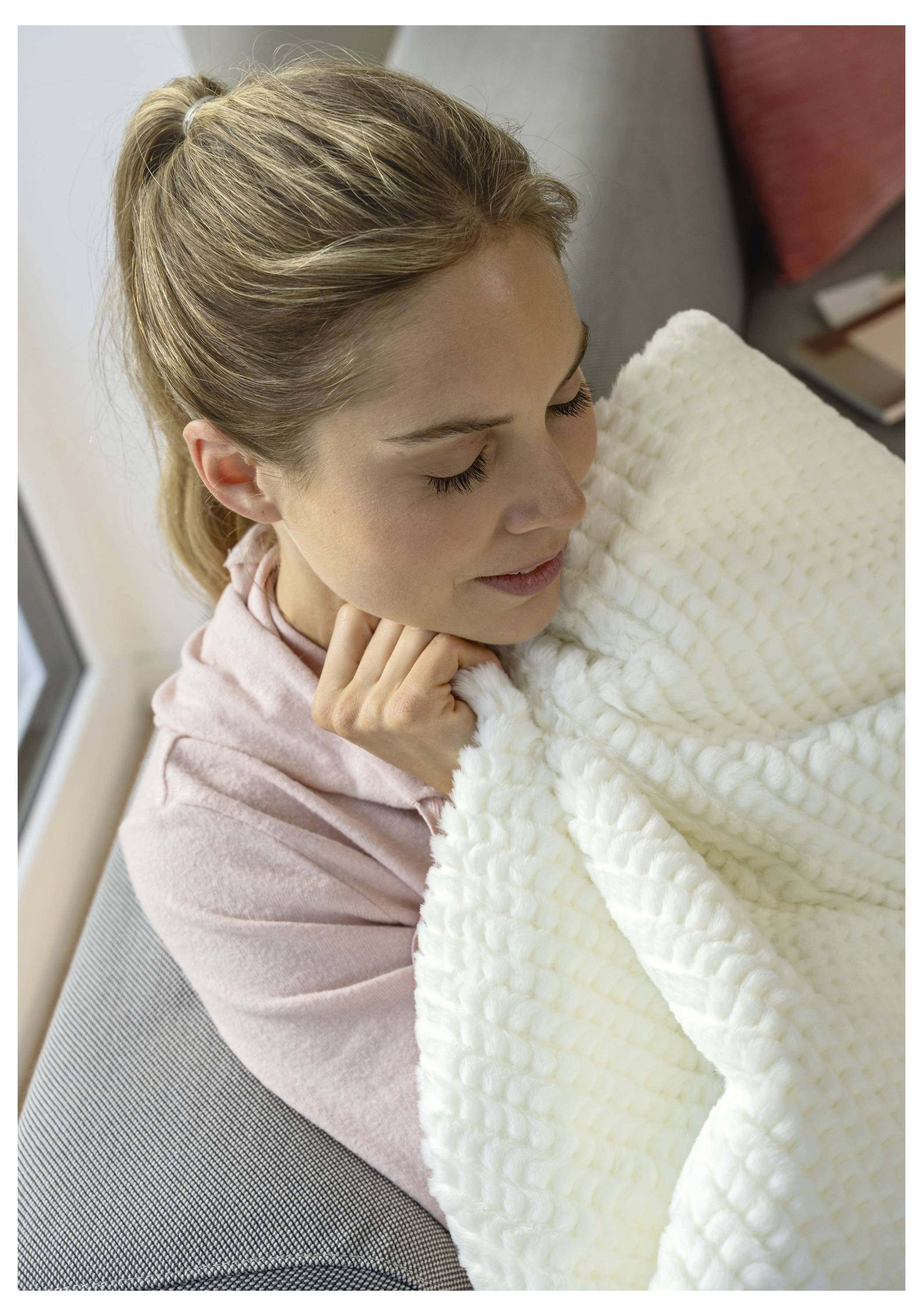 A person gently holds a textured white blanket while seated indoors, wearing a pink top, with a calm expression.
