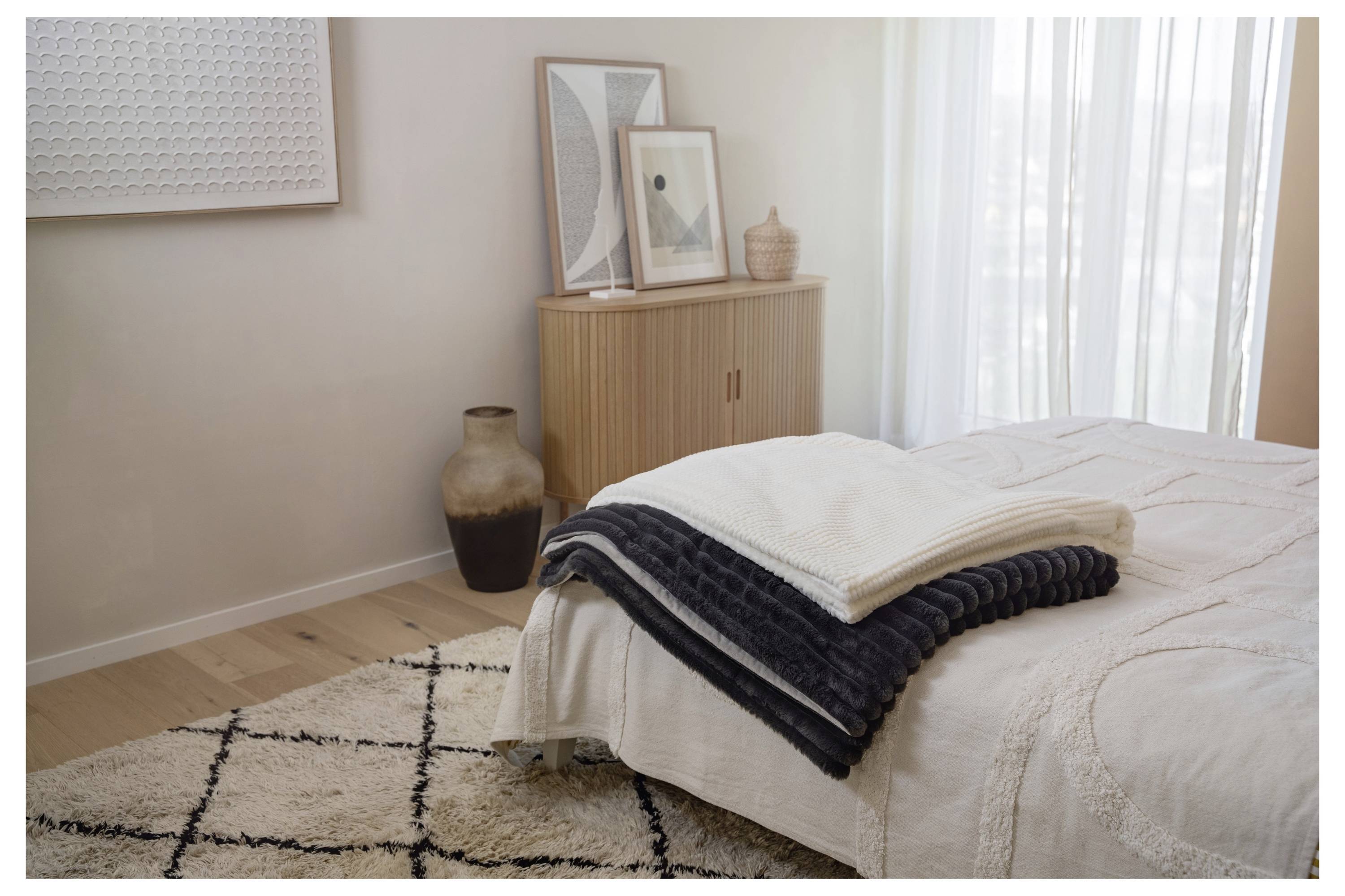 A neatly made bed with gray and white blankets, a patterned rug on wooden floor, a wooden cabinet with framed art and a large vase nearby.