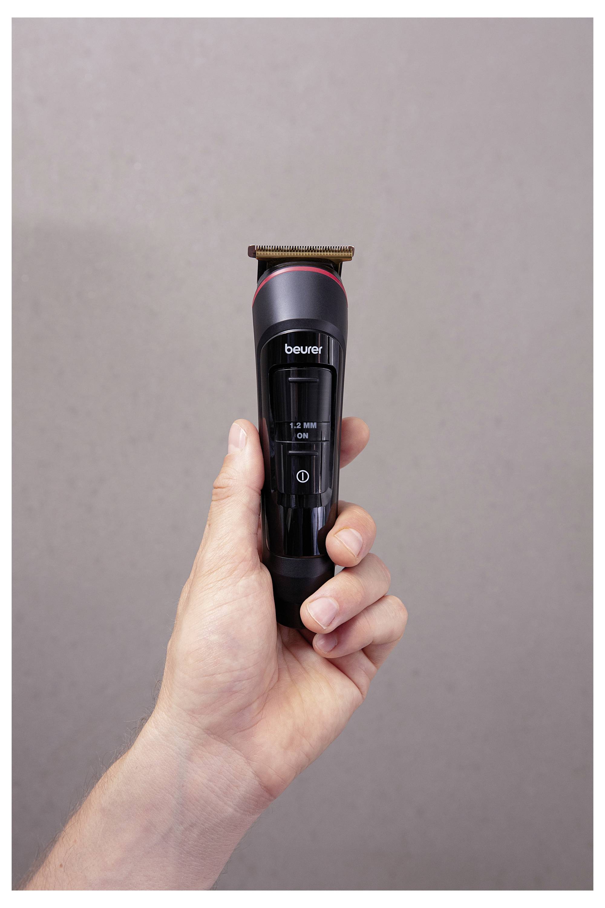 A person holds a black and red electric beard trimmer against a neutral background, showing the brand name and power button detail.
