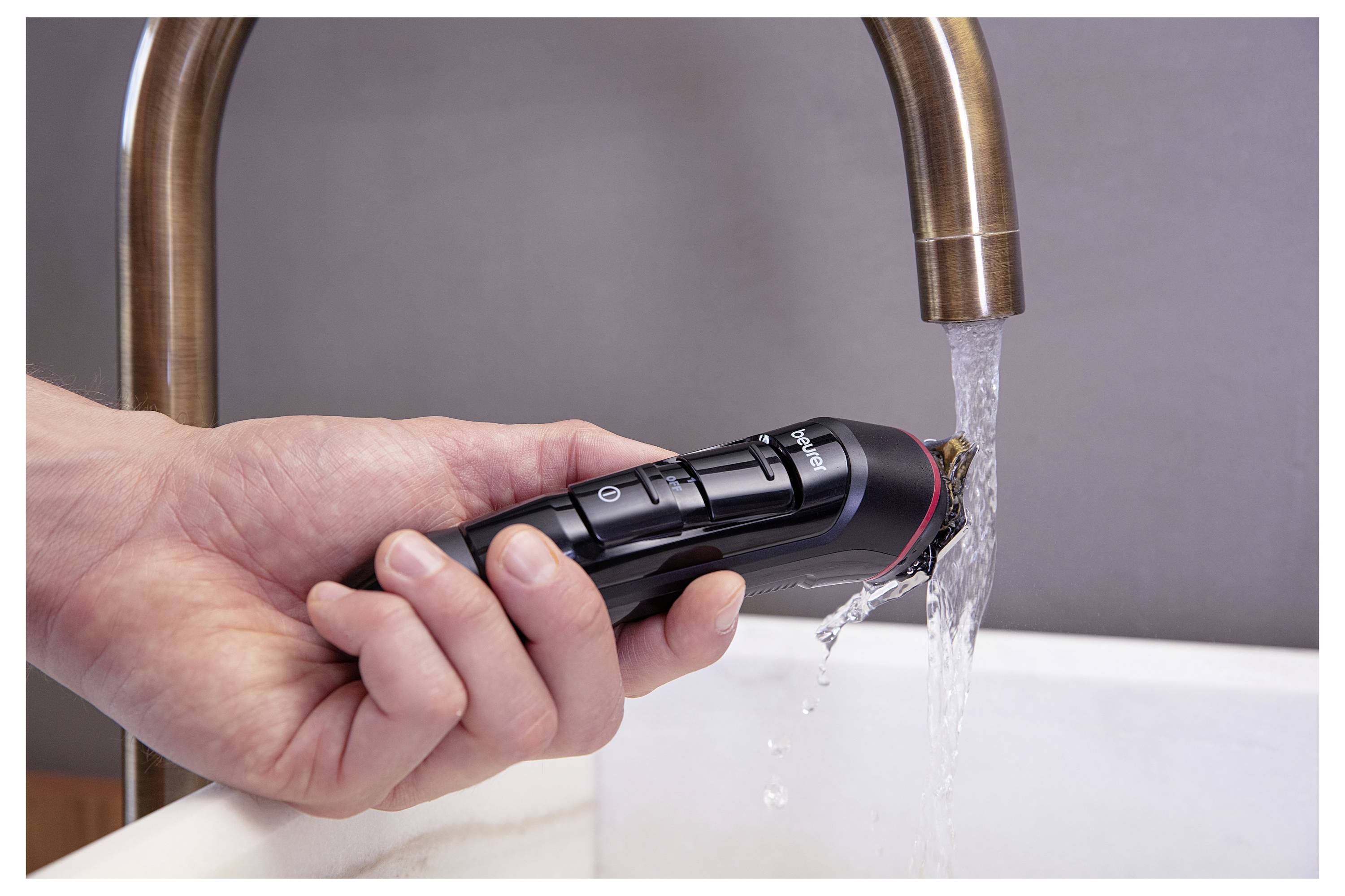 A hand holds a black electric razor under a running faucet in a sink, demonstrating its waterproof feature for cleaning.