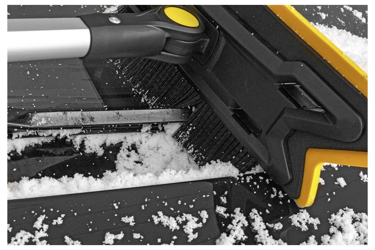 A snow brush with a black bristle head and yellow details is clearing snow from a car windshield.
