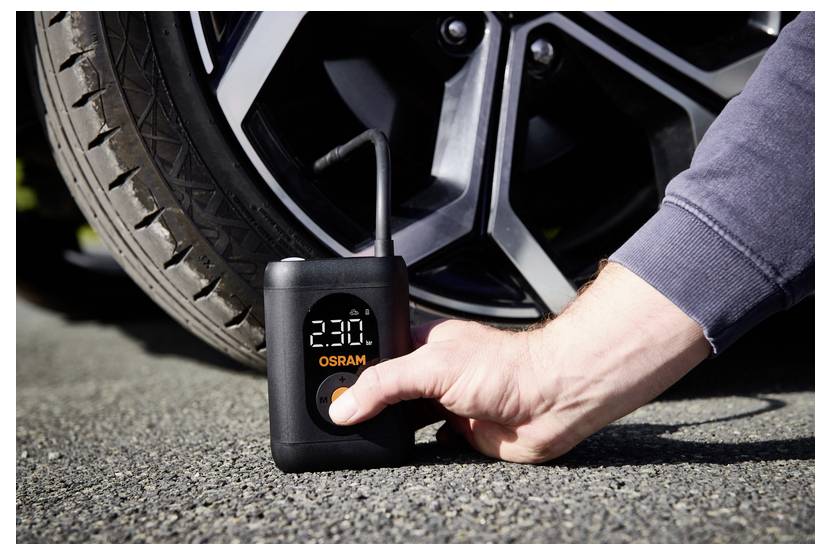 A hand uses a small, portable tire inflator to check the air pressure on a car tire. The digital display shows '23.0'.