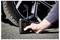 A hand uses a small, portable tire inflator to check the air pressure on a car tire. The digital display shows '23.0'.