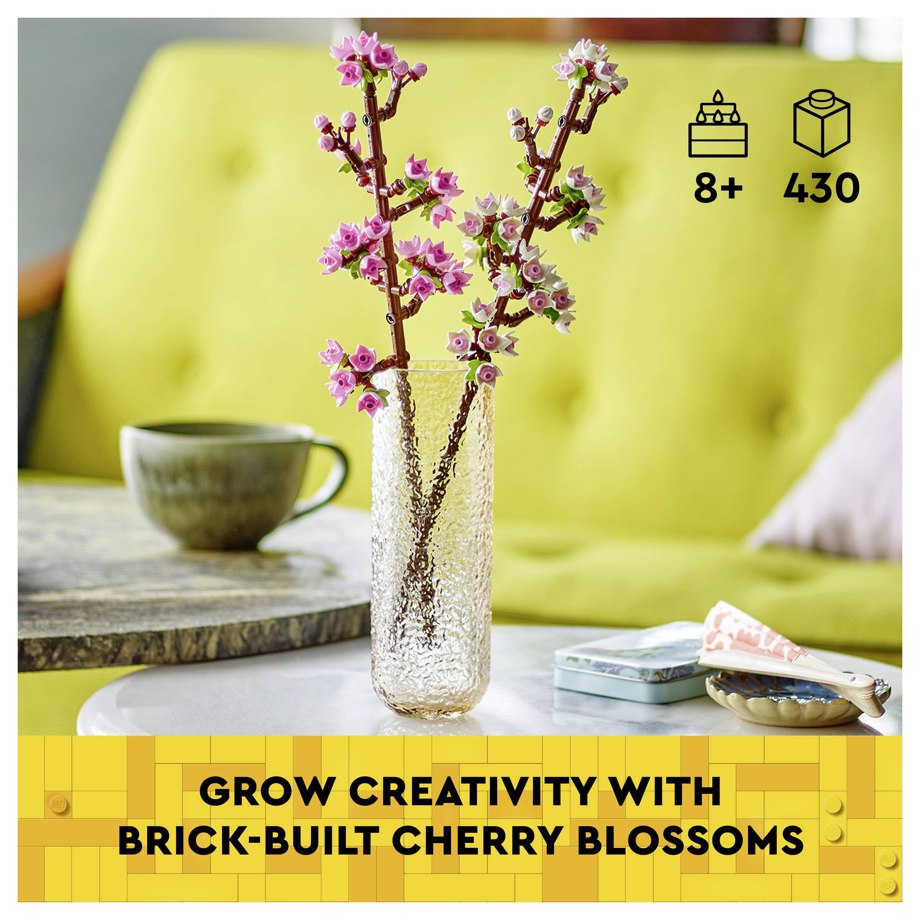 'Grow creativity with brick-built cherry blossoms' text, two pink brick cherry blossom branches in a vase on a table with tea and cookies.