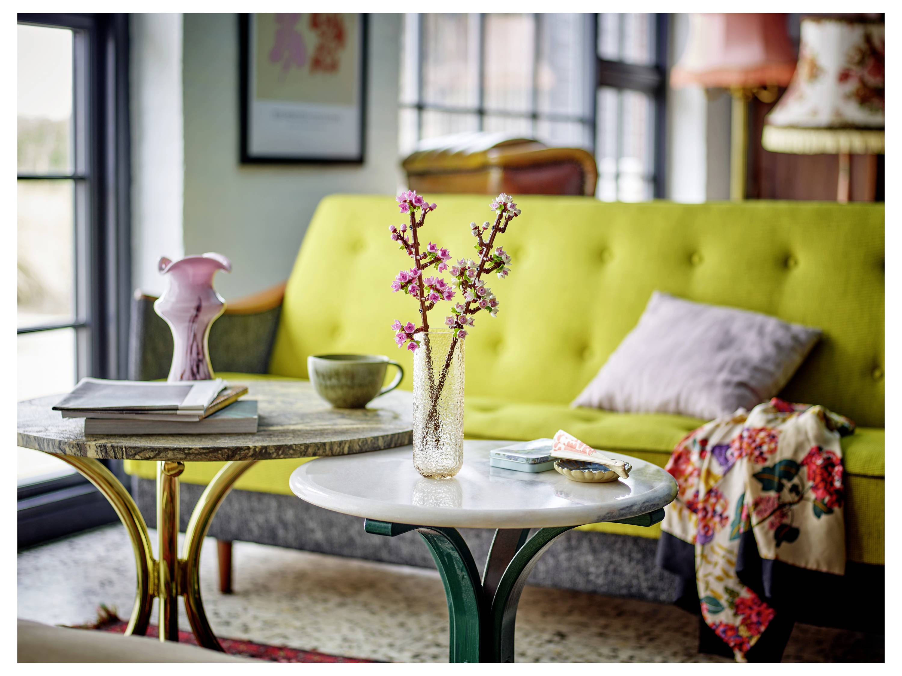 A vase with cherry blossoms on a small table, set in a living room with a yellow sofa, a pink pillow, and decorative objects.