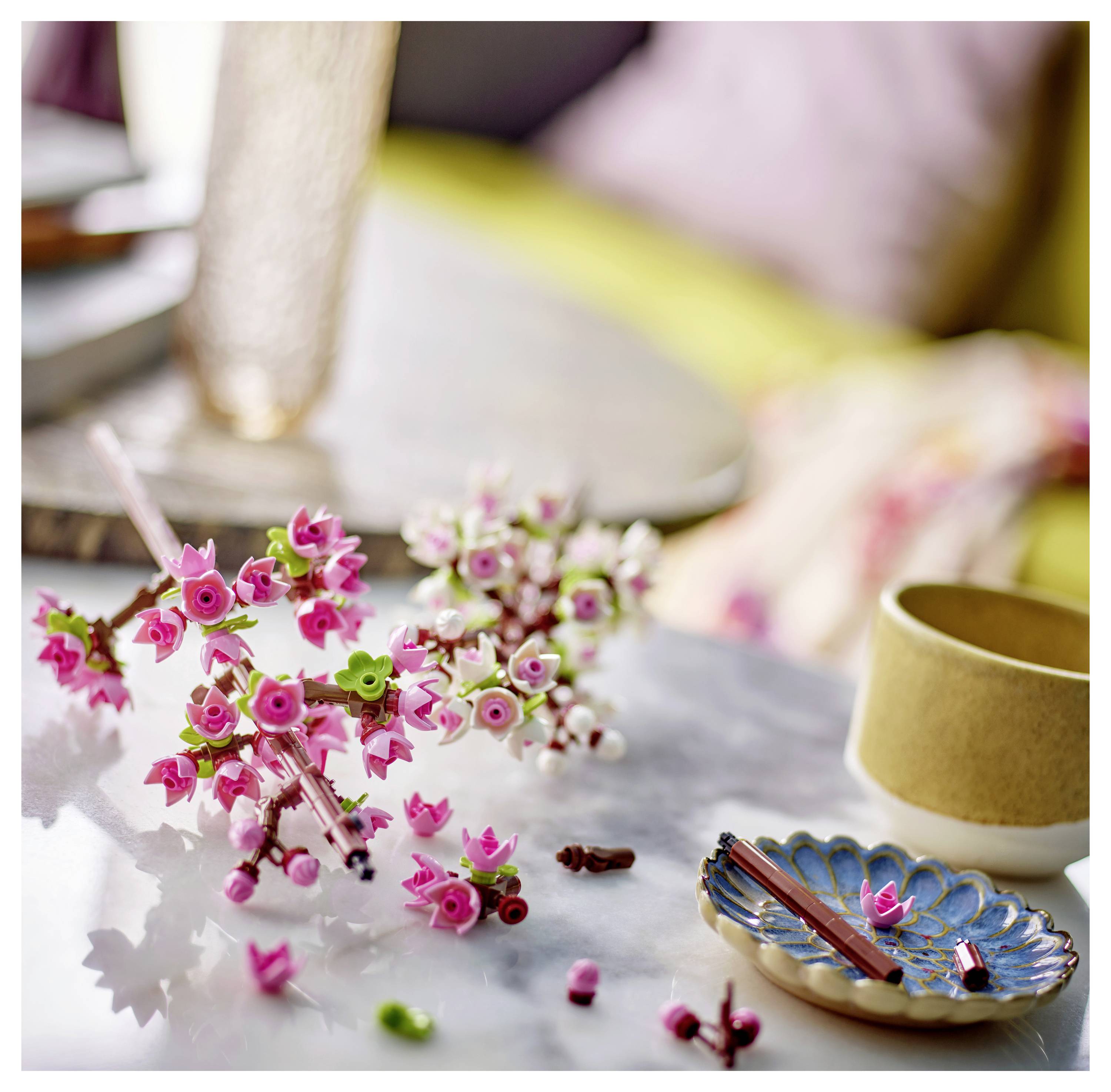 A close-up of small, pink and green artificial flower decorations on a marble surface, beside a beige cup and a decorative plate.
