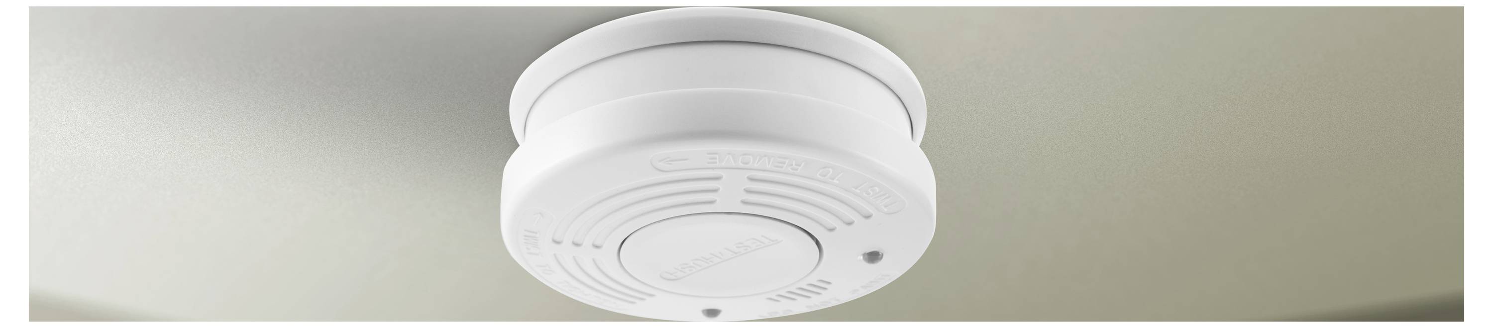 A ceiling-mounted smoke detector with a white circular design is installed on a beige ceiling, indicating a safety device in use.