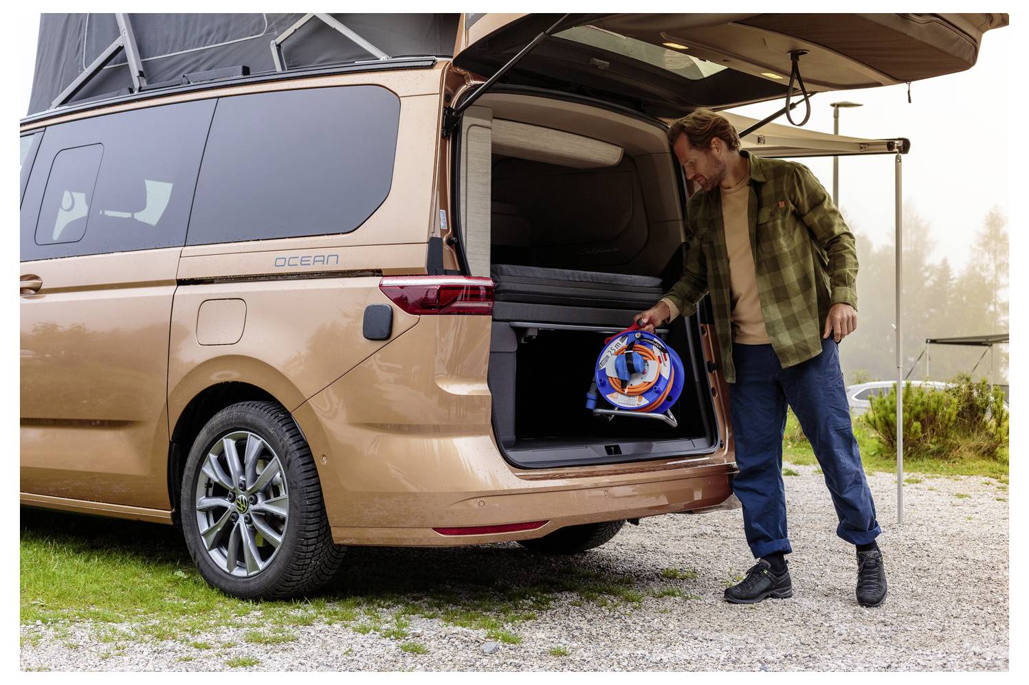 A person placing camping gear in the trunk of a bronze van parked on a gravel area surrounded by greenery.