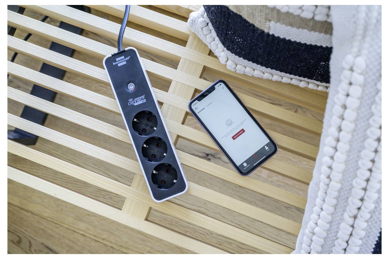 'Phone with an app open next to a power strip on a wooden bench, partially covered by a textured blanket, suggesting home automation.'