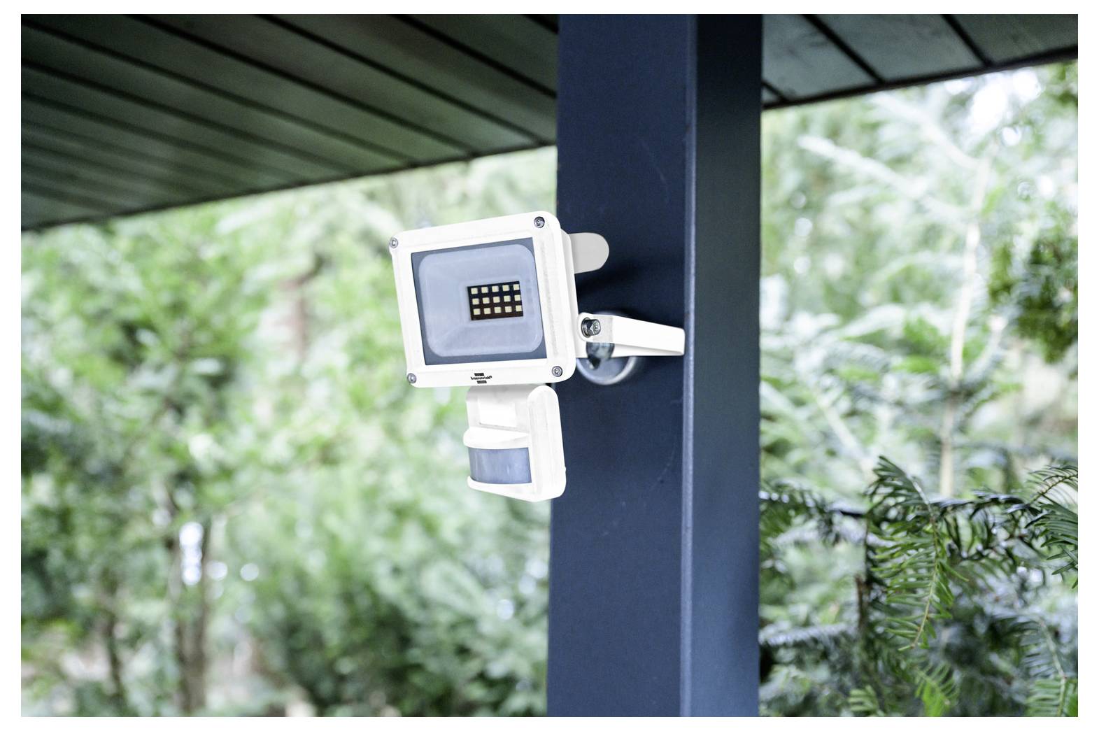An outdoor security camera attached to a post under a roof, surrounded by greenery, focused on monitoring the surrounding area.