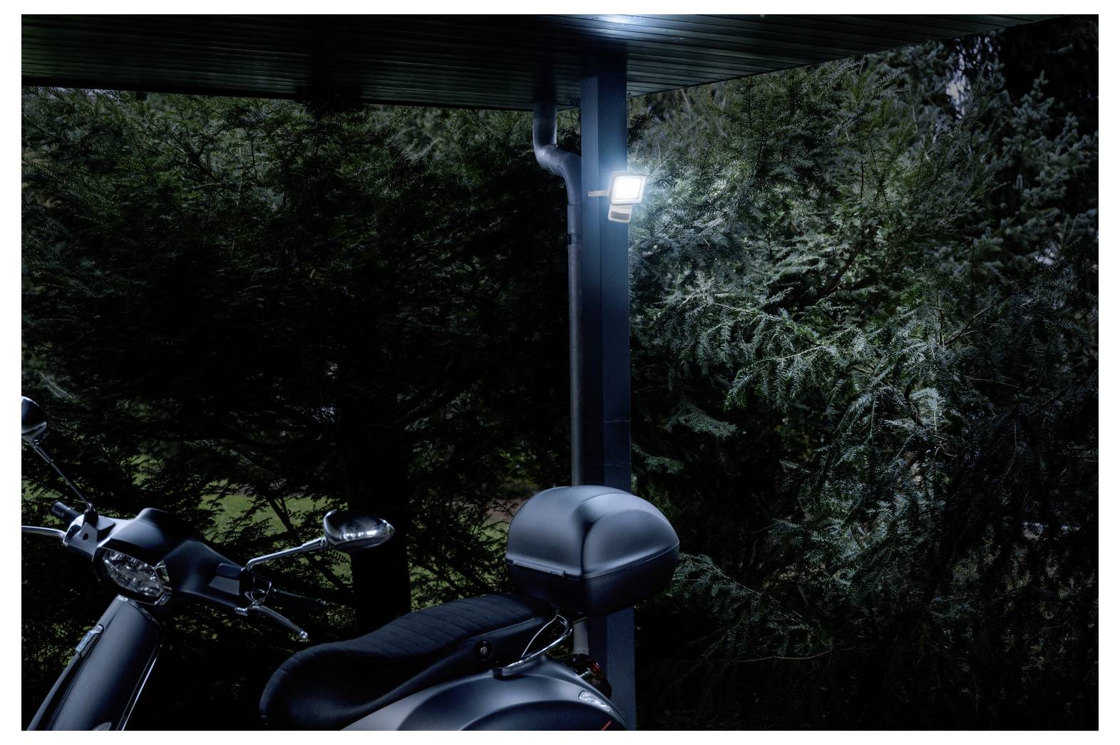 'A motorcycle is parked under a roofed area, next to a pole with an illuminated light. Trees and foliage are visible in the background.'