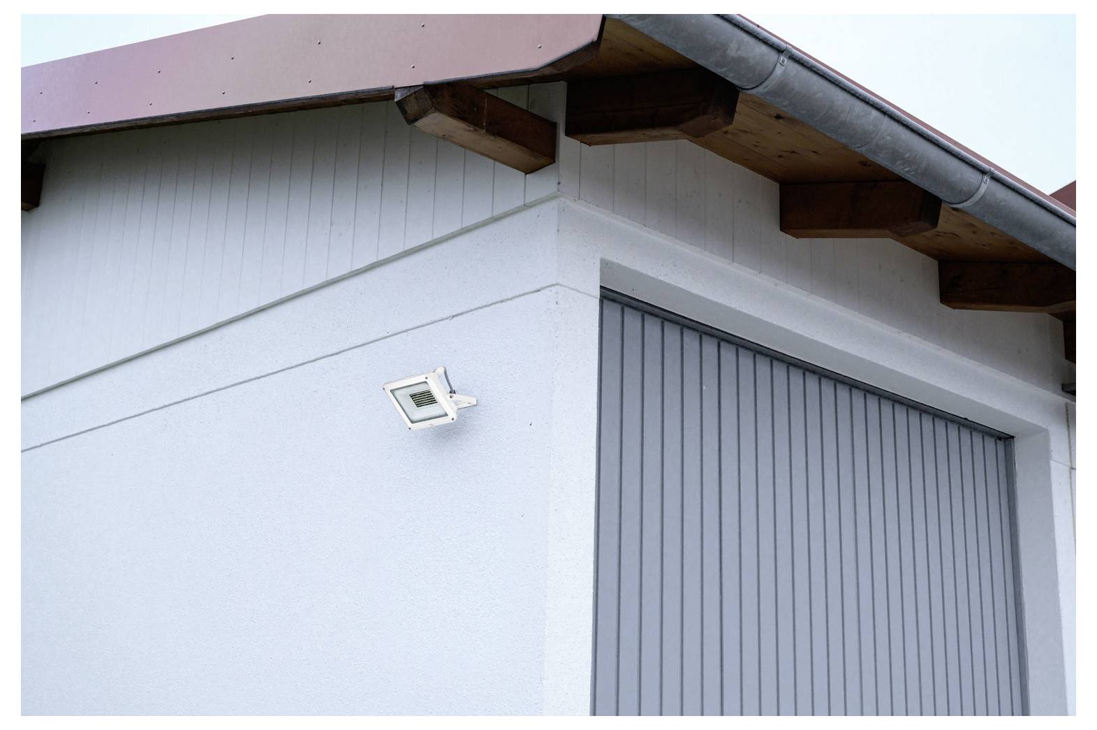 A white building corner with a pitched roof features a mounted security camera on its wall, near a large, closed, vertical gray garage door.