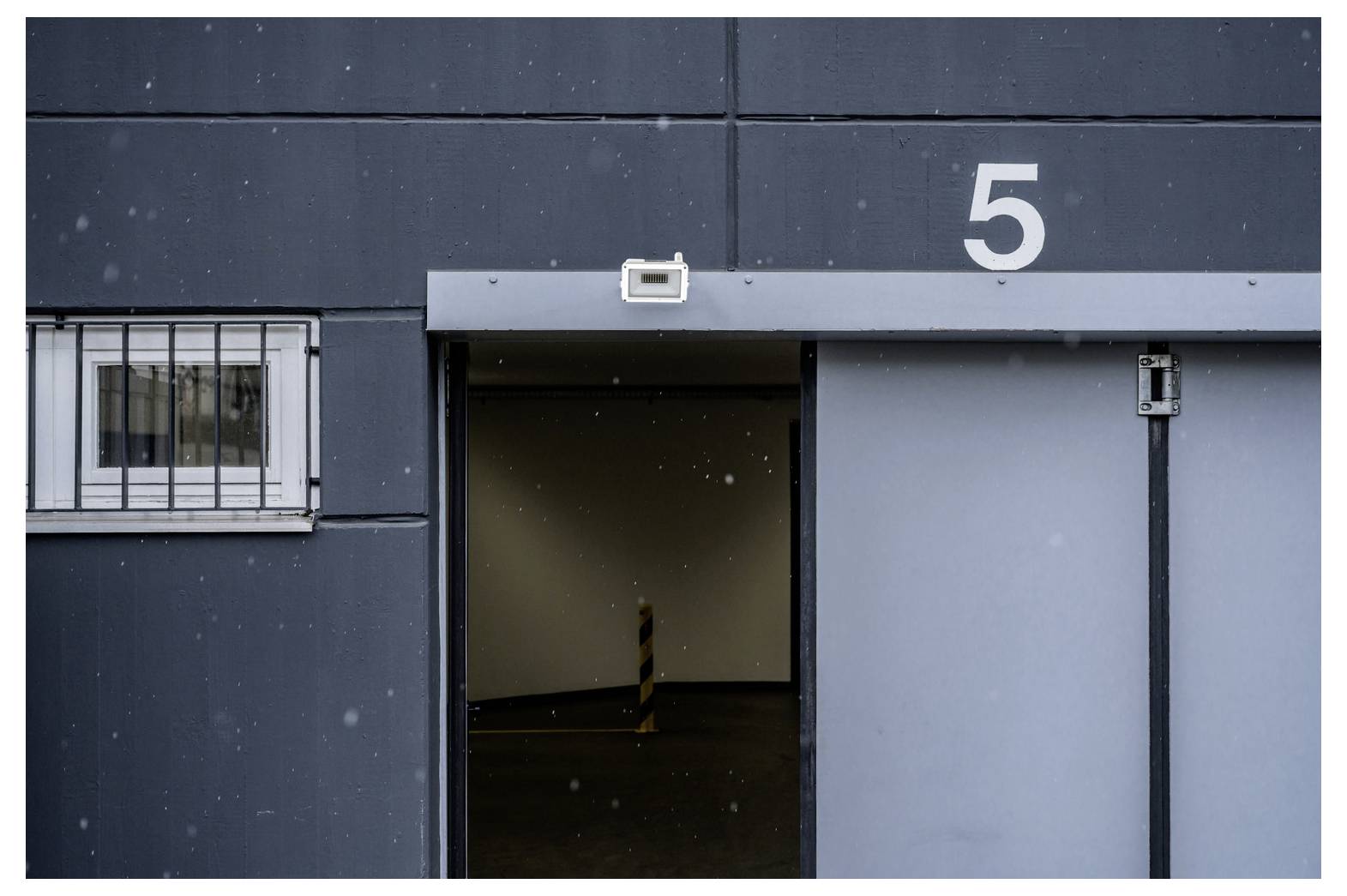'Gray industrial building exterior with an open garage door labeled 5, leading to a dimly lit interior. A barred window is on the left.'