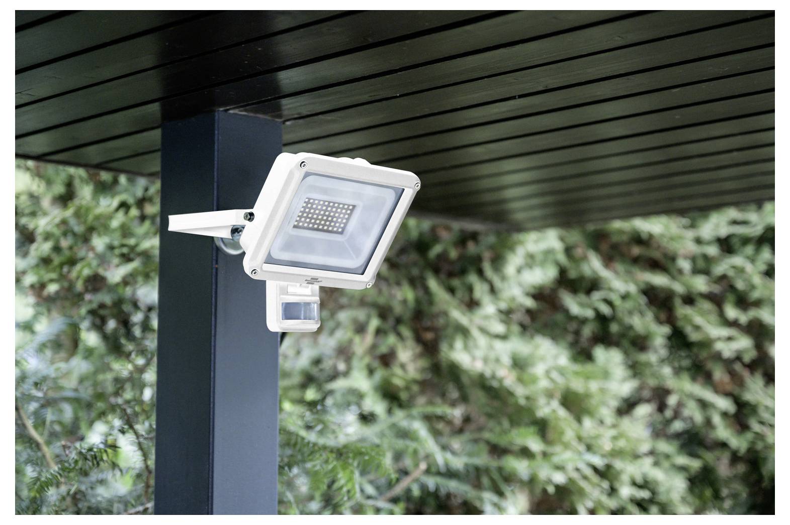 Outdoor floodlight with motion sensor mounted on a patio roof support, surrounded by green foliage.