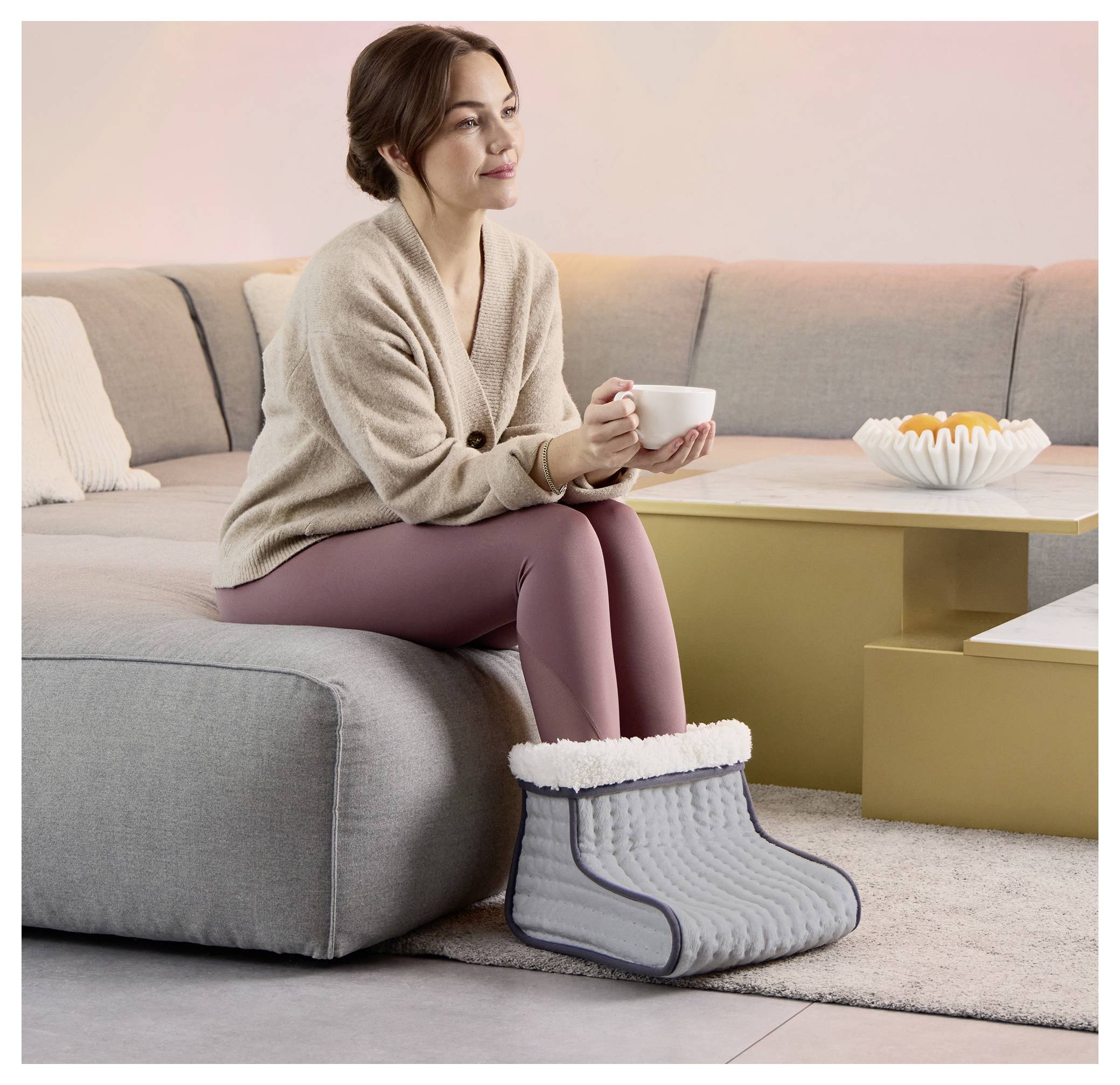 A woman sitting on a sofa holds a cup, with her feet in a gray, fabric foot warmer. A modern living room setting with a beige sofa.