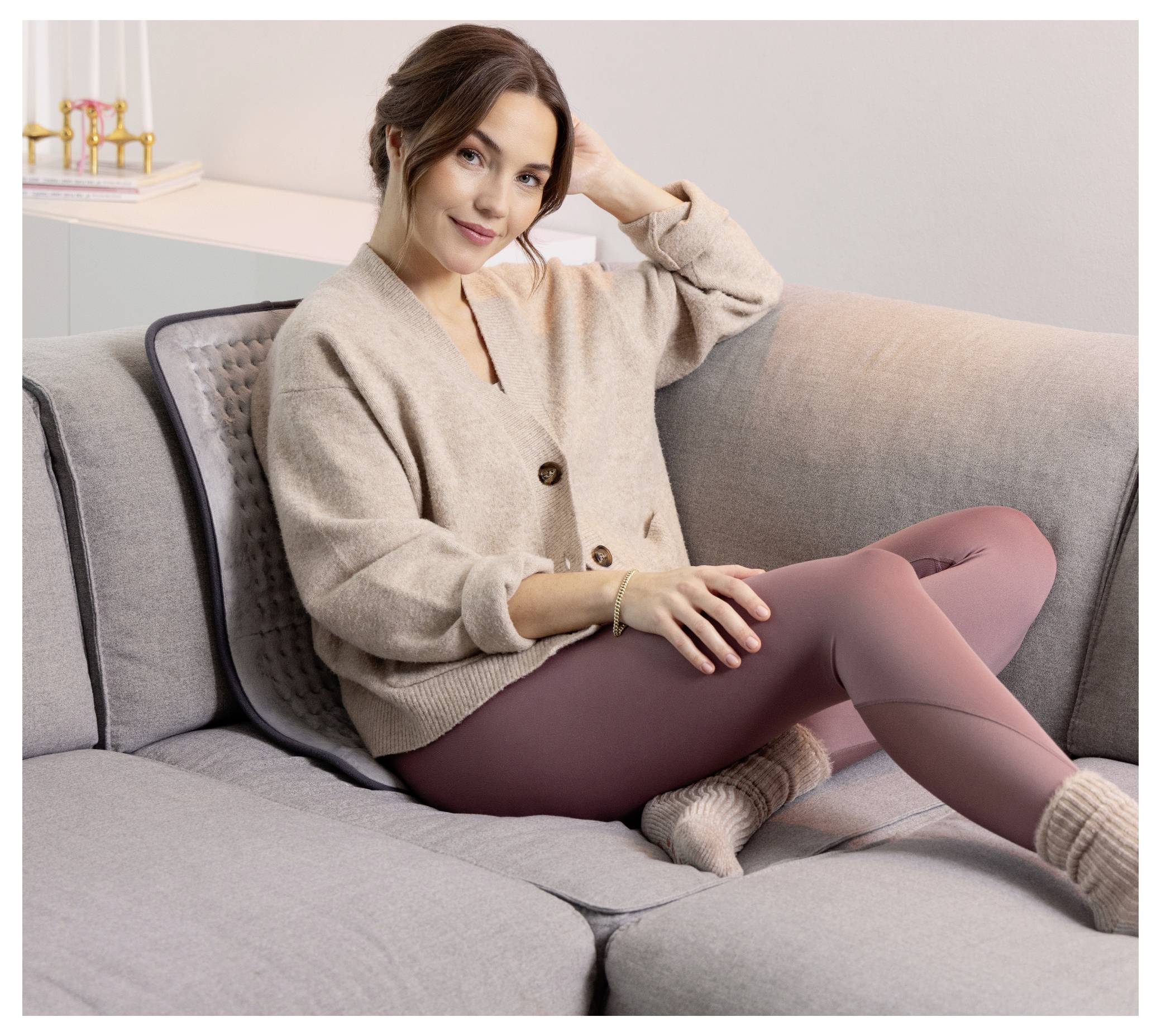 A person sitting comfortably on a grey couch, wearing a beige sweater and mauve leggings, with a relaxed expression, in a cozy living room.
