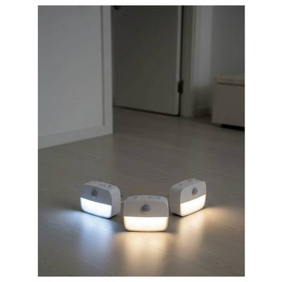 Three rectangular nightlights with motion sensors on a wooden floor emit blue, white, and warm orange light in a dimly lit room.