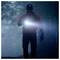 A person in outdoor gear walks through a misty forest at night, holding a bright flashlight that illuminates their path.