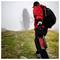 A person in a red jacket carrying survival gear walks toward a large satellite dish in a foggy field, suggesting exploration or research.