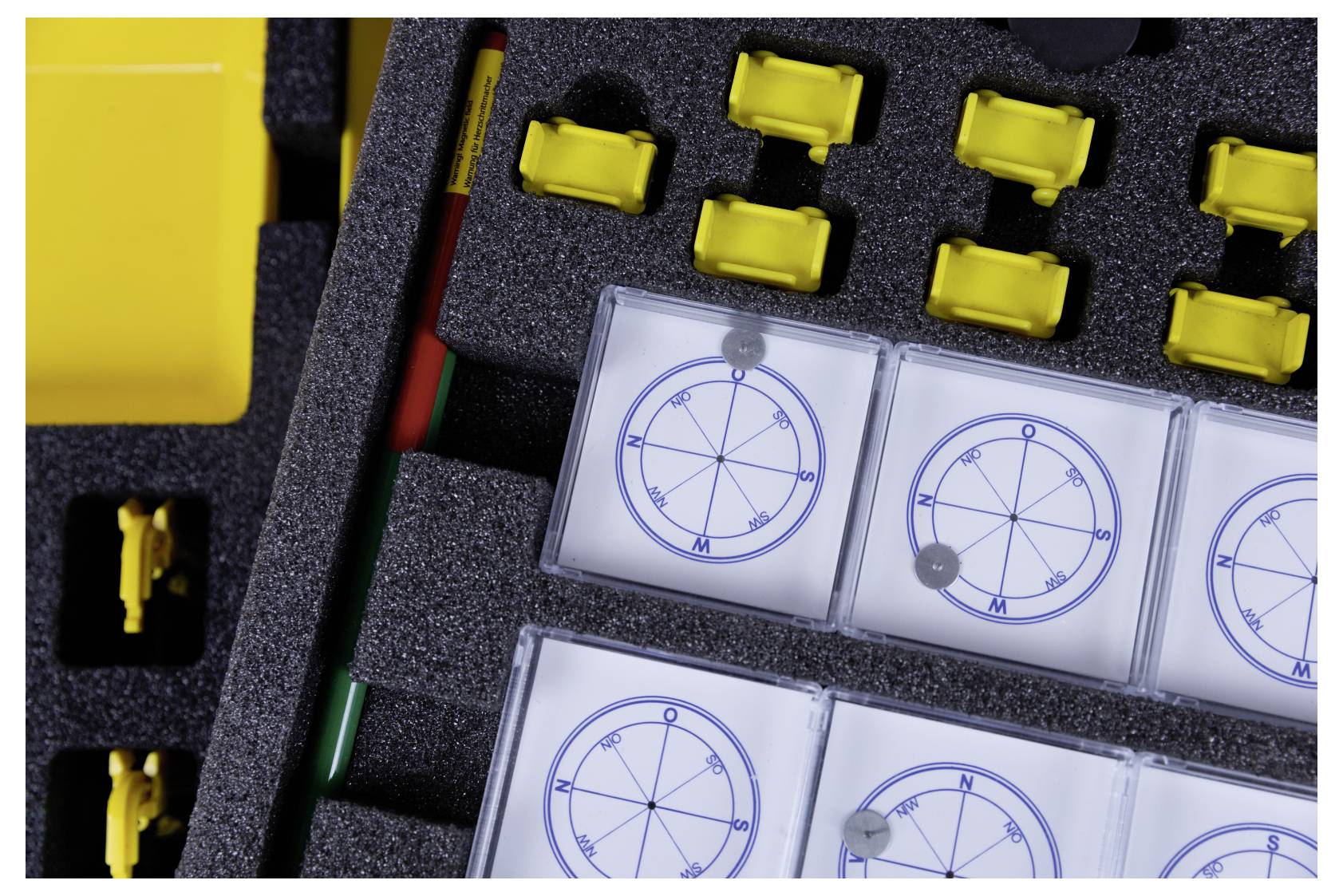 A foam case holds several yellow plastic fittings, two rods, and clear compasses with cardinal directions, indicating navigational tools.