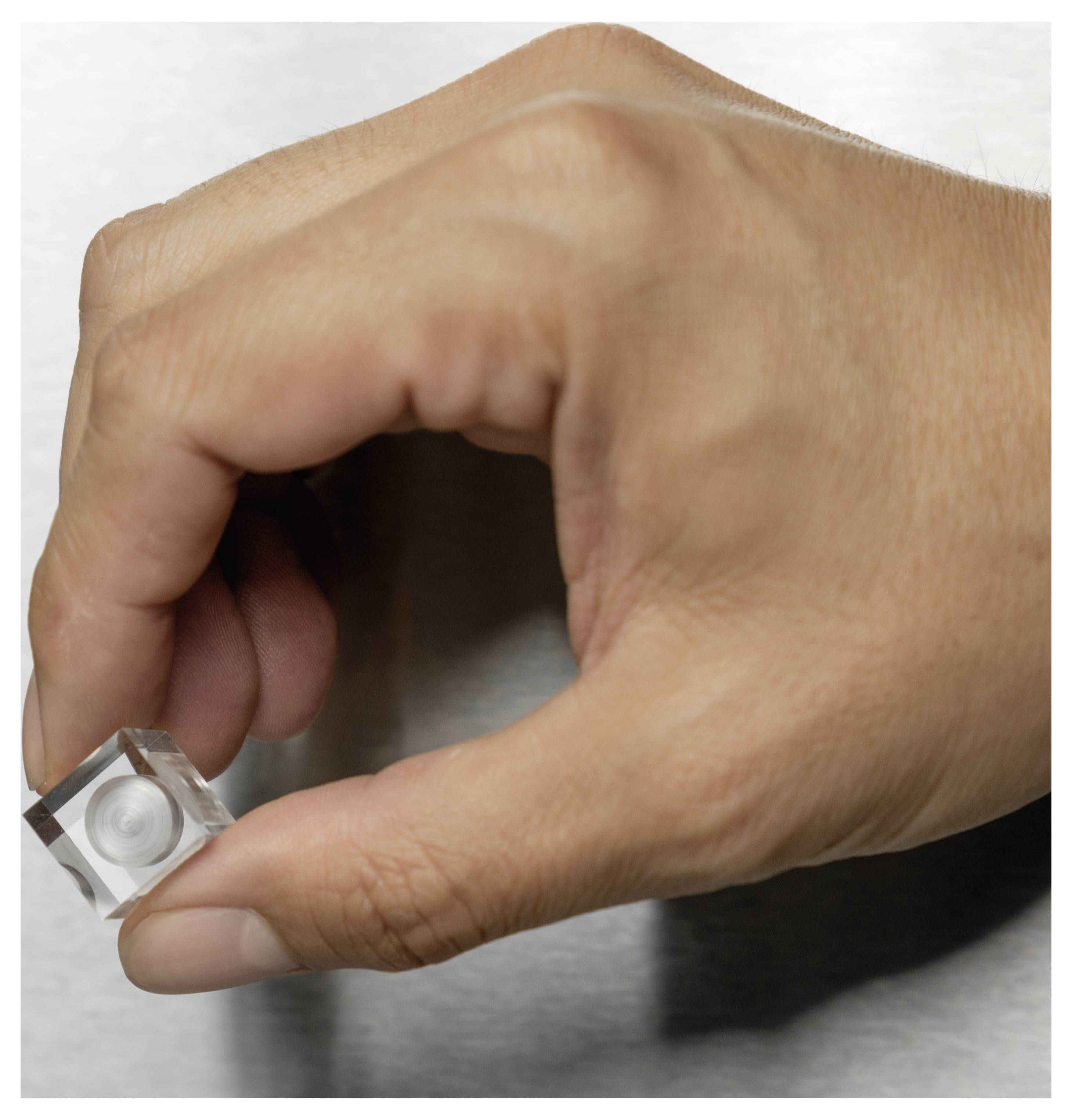 A hand holding a small, transparent cube with a circular indentation, positioned against a plain background.