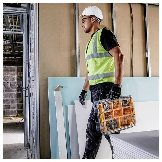 A construction worker in a high-visibility vest and hard hat carries a toolbox in a partially-built room, indicating ongoing construction work.