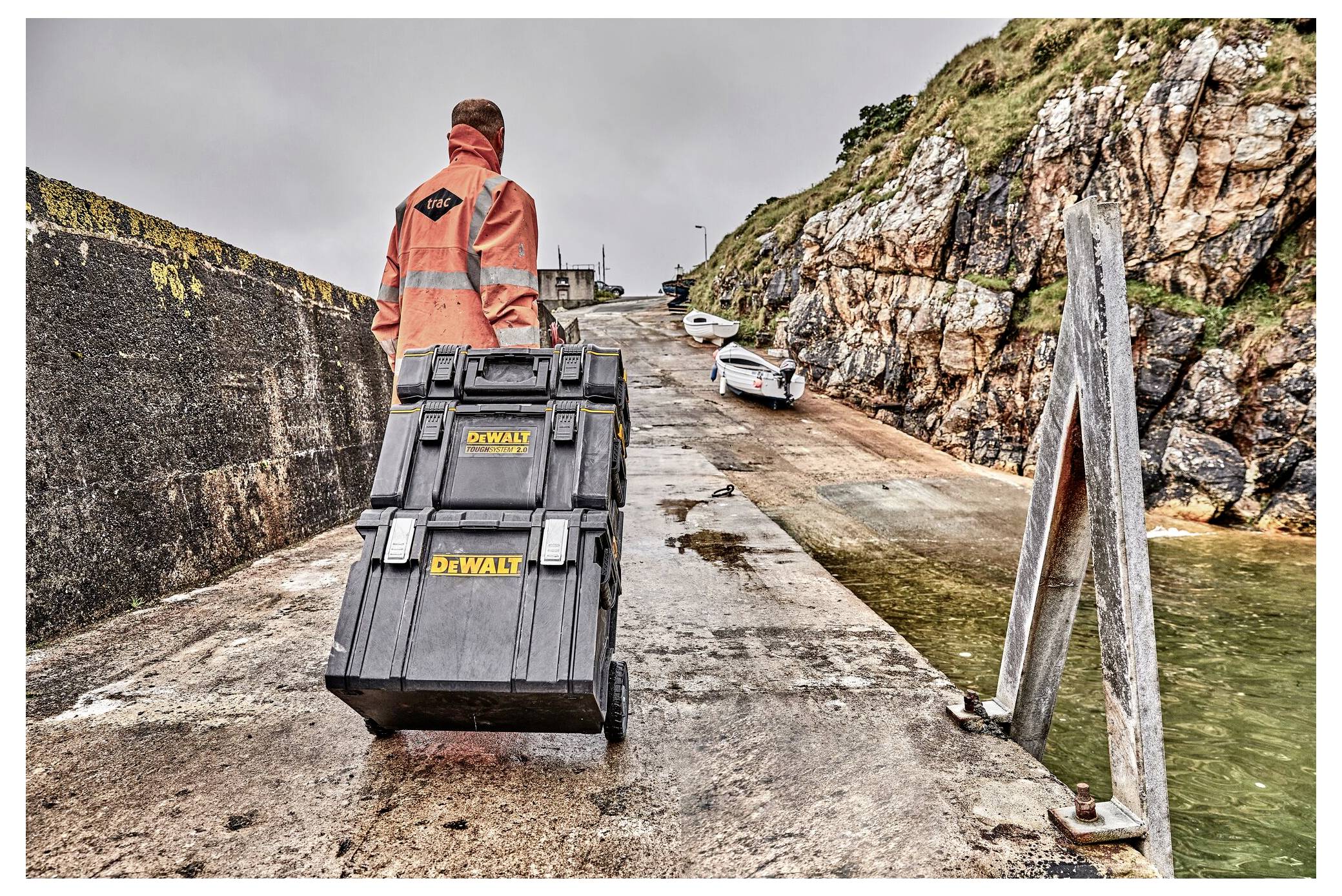 A person in an orange jacket pulls a tool chest on wheels up a concrete ramp near a rocky shoreline, with boats visible in the distance.