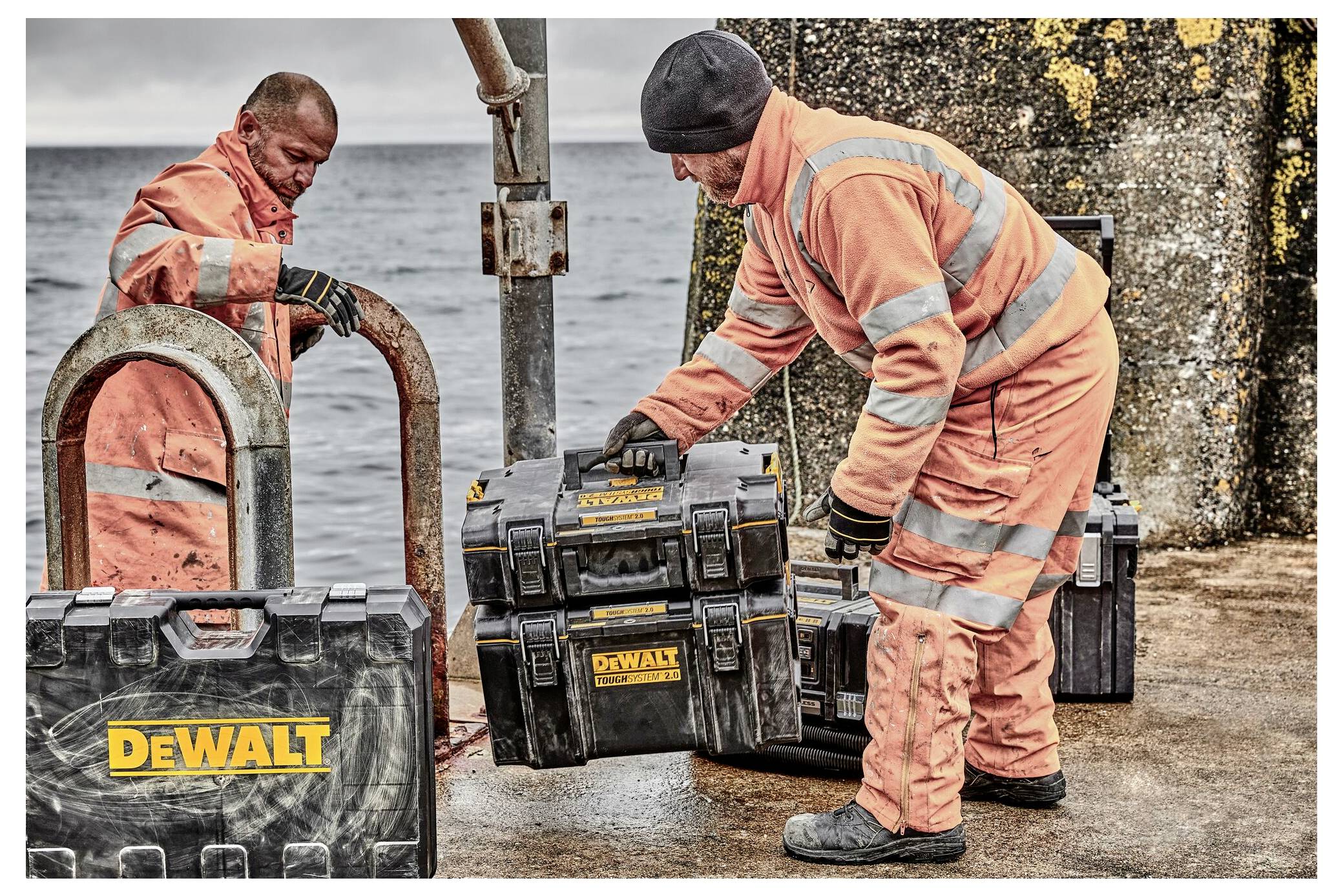 Two workers in orange reflective clothing handle black and yellow toolboxes labeled 'DeWalt' on a pier with the sea in the background.