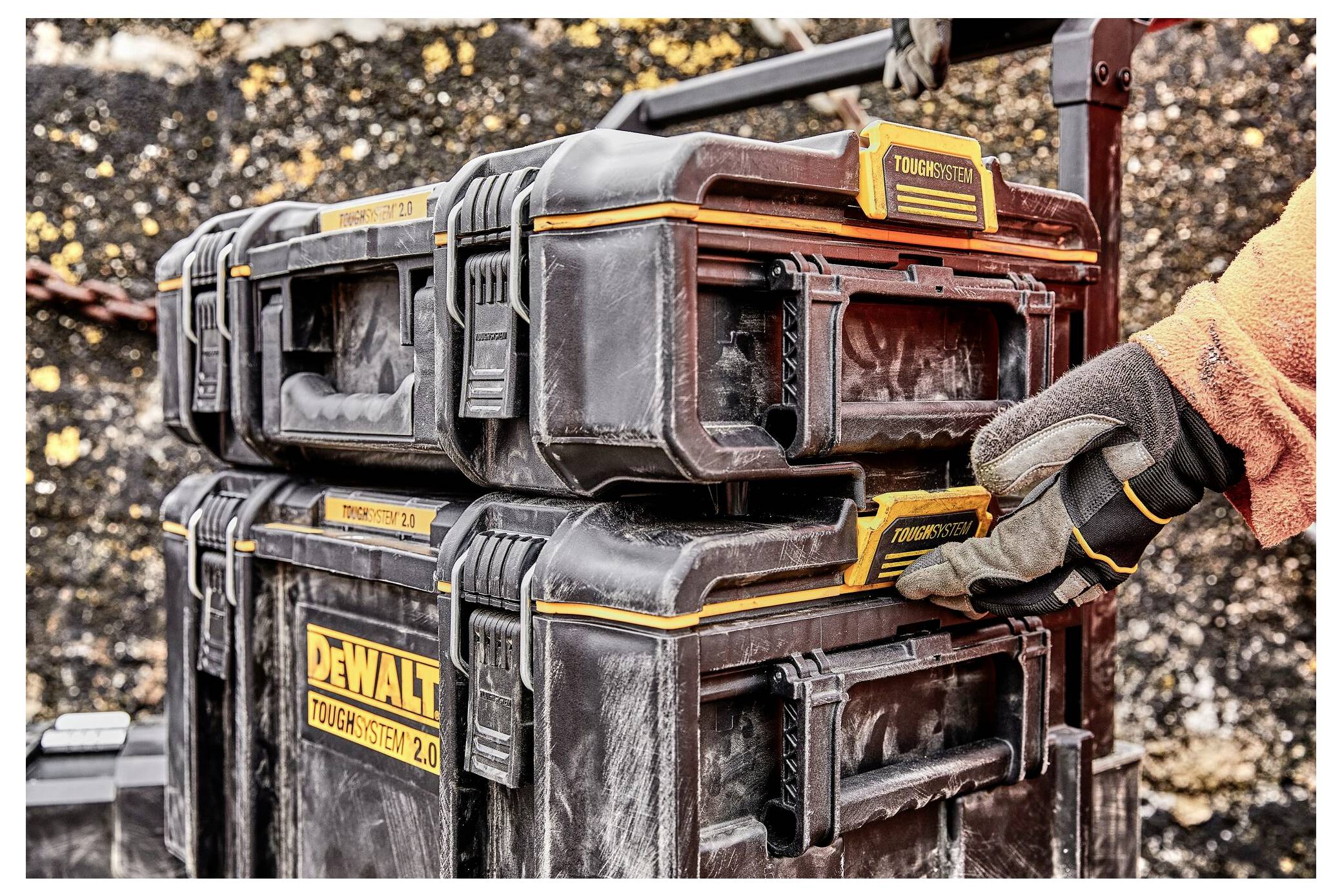 A gloved hand grips the latch of a stacked, rugged black toolbox. The label reads 'TOUGHTSYSTEM 2.0'. The background shows a gritty, outdoor setting.
