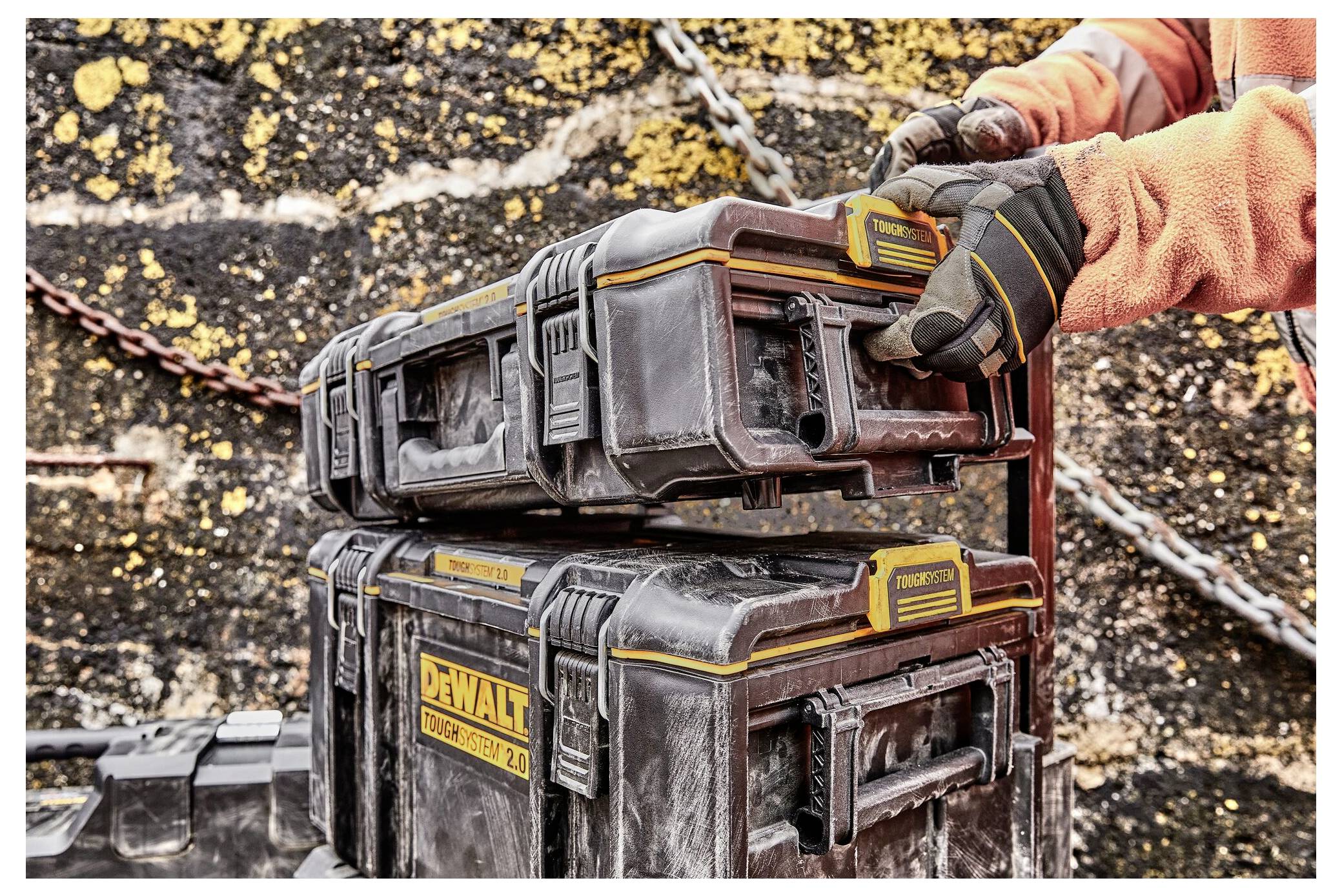 A person wearing gloves and an orange jacket handles two stacked DeWalt toolboxes, highlighting rugged outdoor use.
